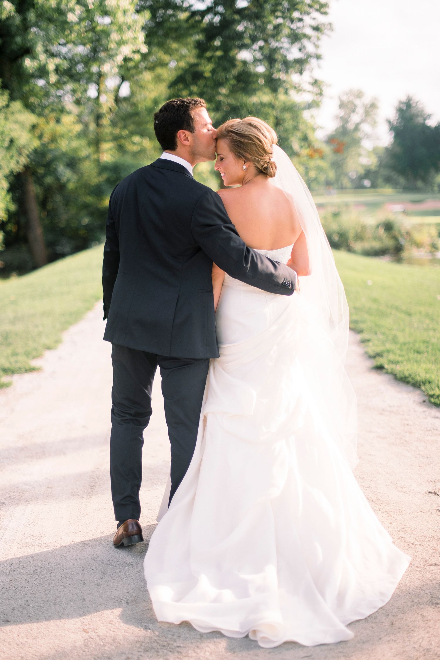 Romantic wedding couple walking and kissing on a sunlit garden path in elegant attire.