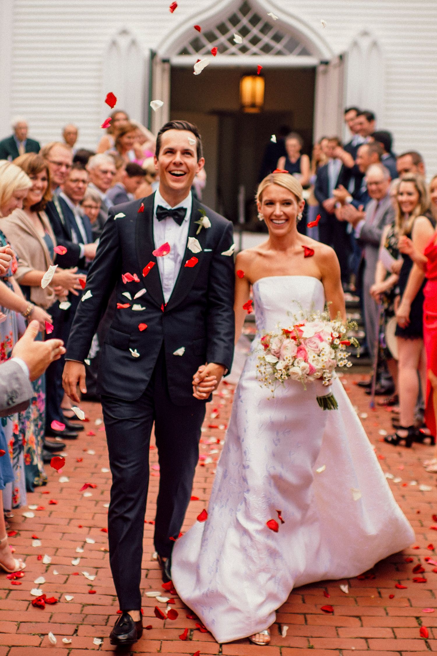 Wedding couple exits ceremony while guests throw rose petals on brick pathway.