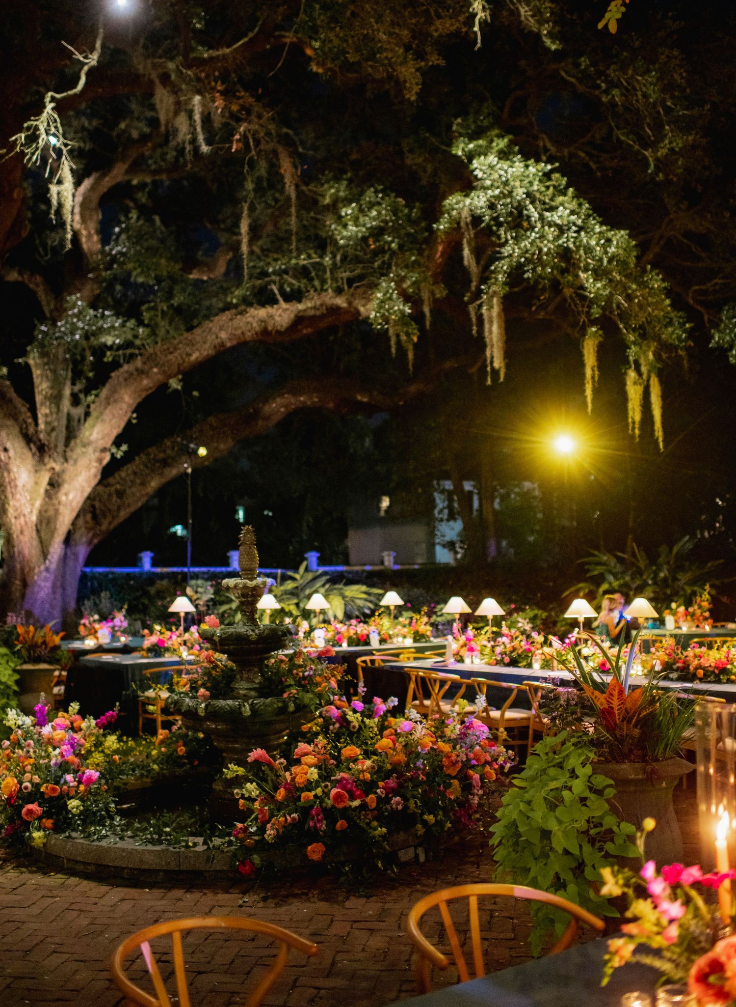 Evening garden reception with illuminated tables and colorful floral centerpieces under oak trees.