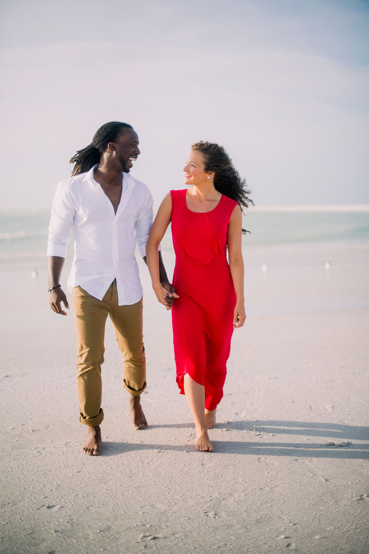 A couple in white and red attire walking together on a sandy beach during a romantic photoshoot.