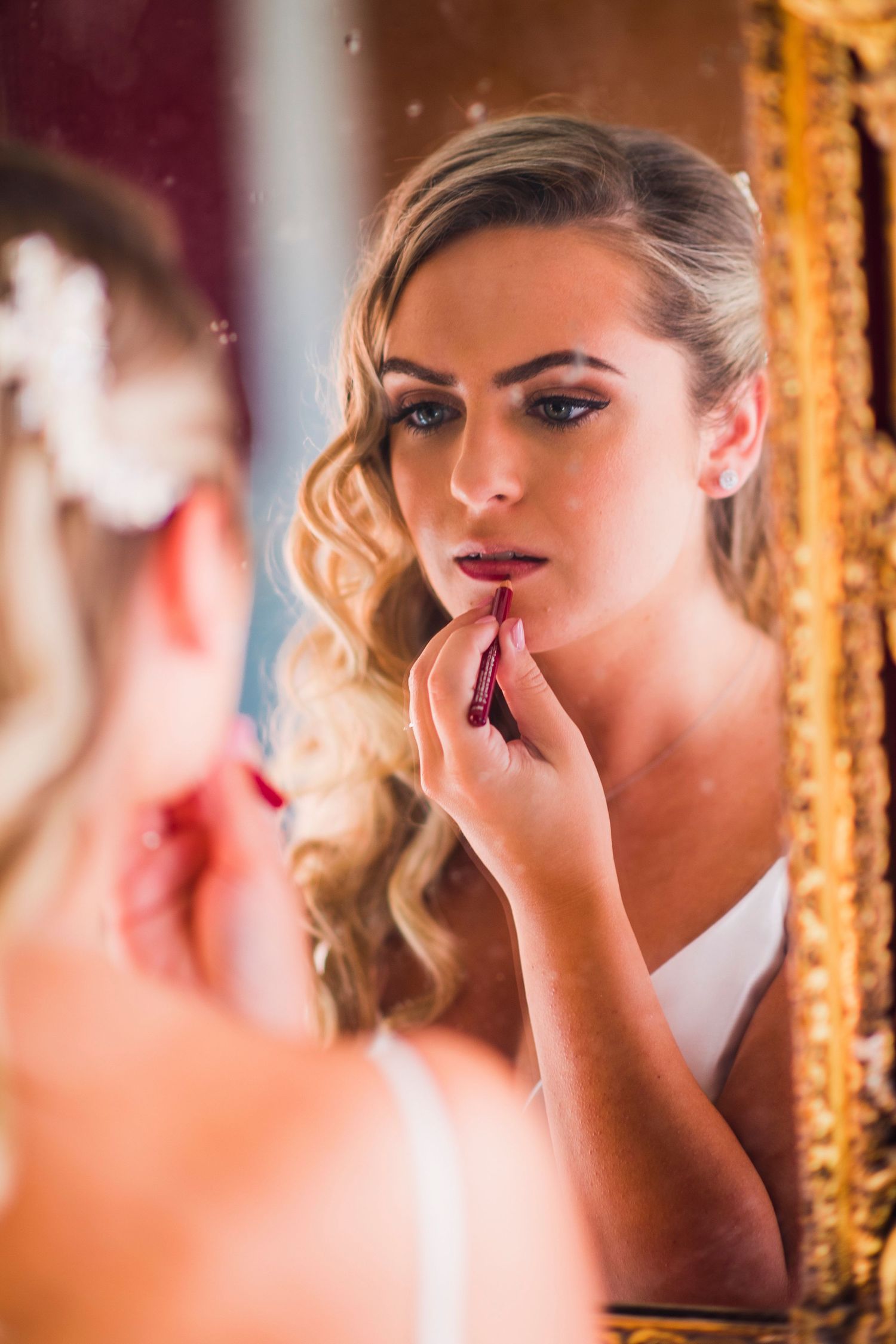 A reflection in an ornate mirror shows someone applying lipstick during wedding preparations.
