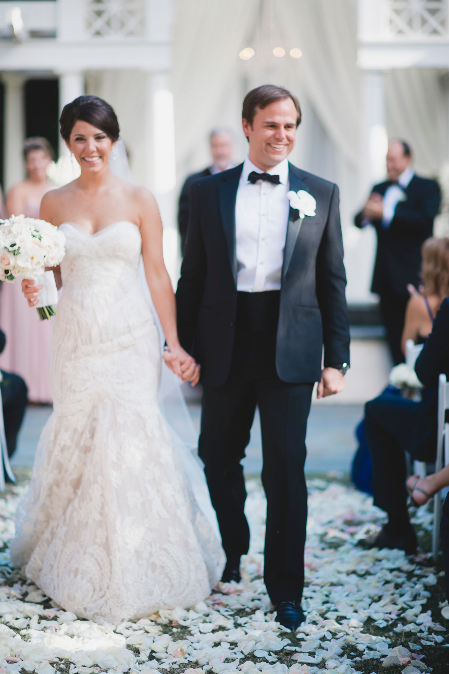 Wedding couple walking through scattered flower petals on elegant white porch.