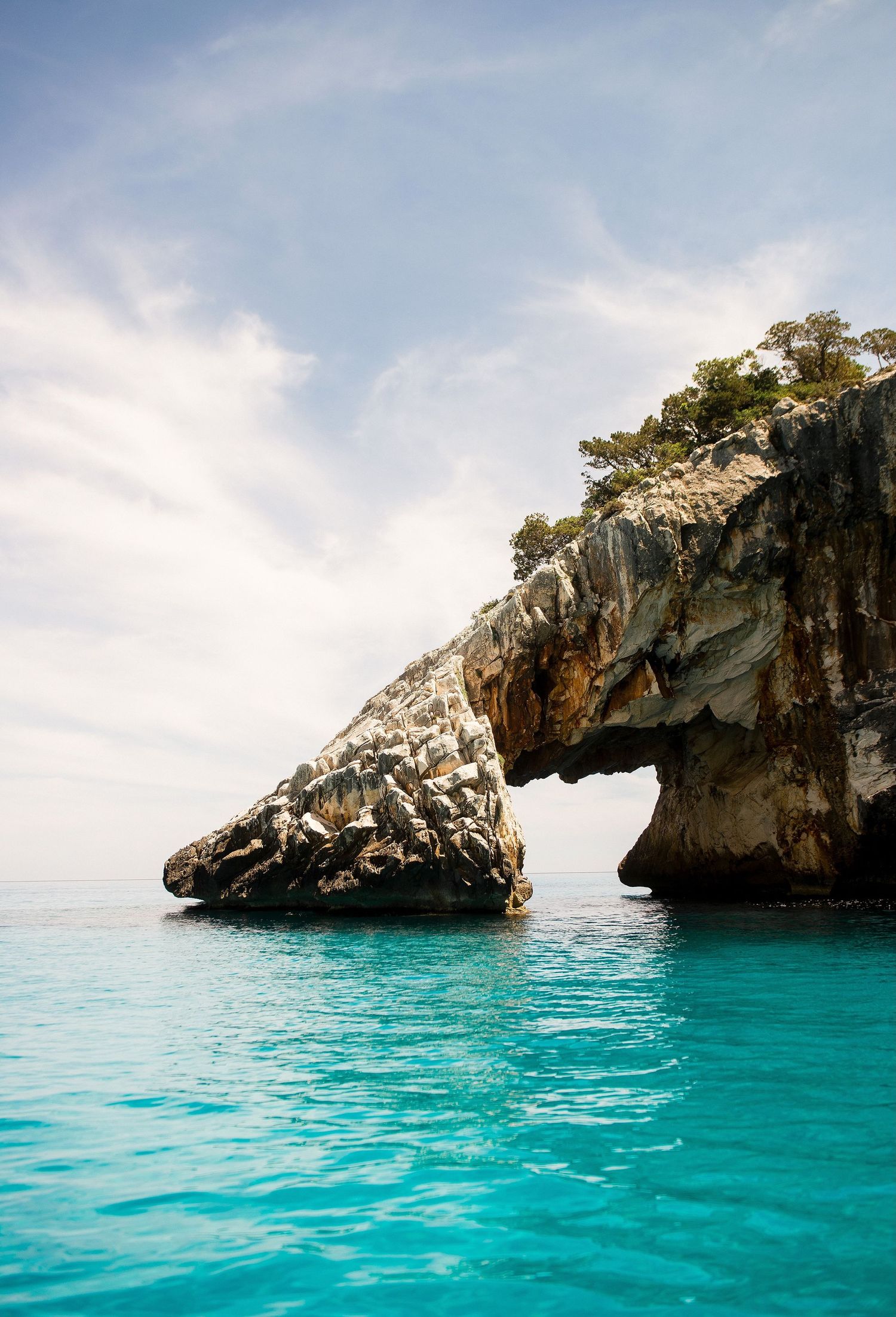 Rugged coastal rock formations rise from crystal clear turquoise Mediterranean waters under bright blue skies.