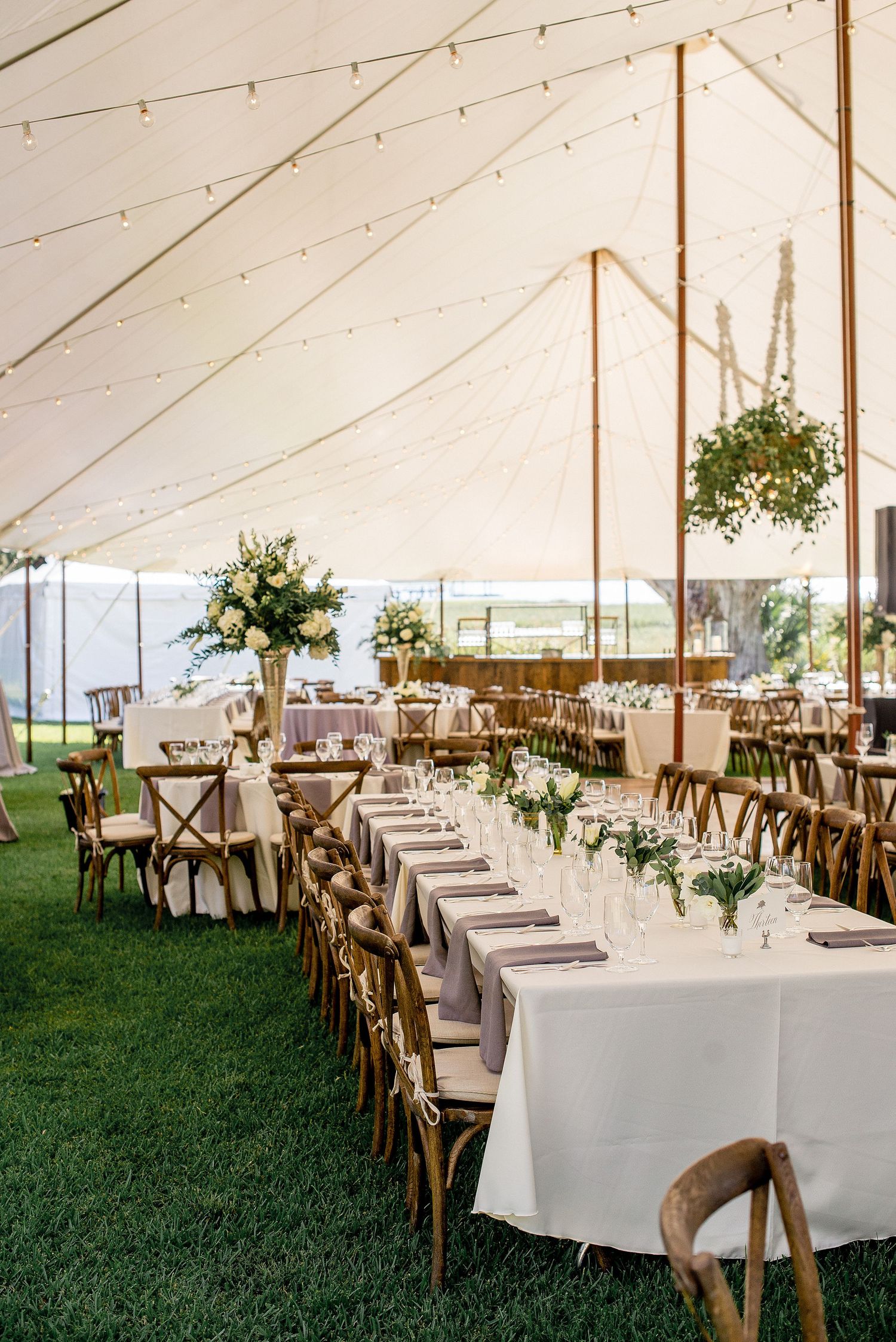 Elegant wedding reception setup under white tent with wooden cross-back chairs and long tables decorated with greenery.