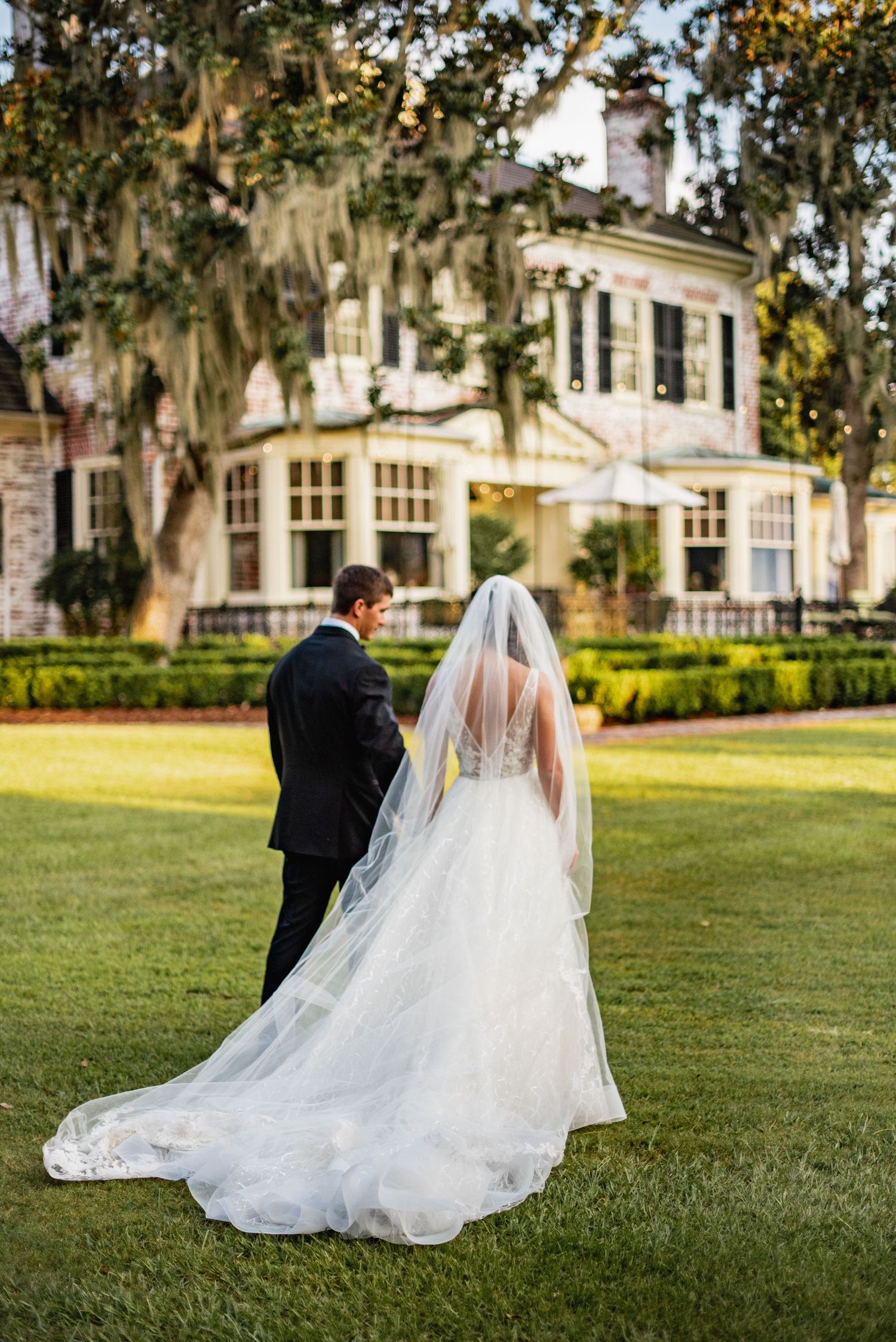 A wedding couple stands on a manicured lawn in front of a grand southern mansion with Spanish moss draped trees.