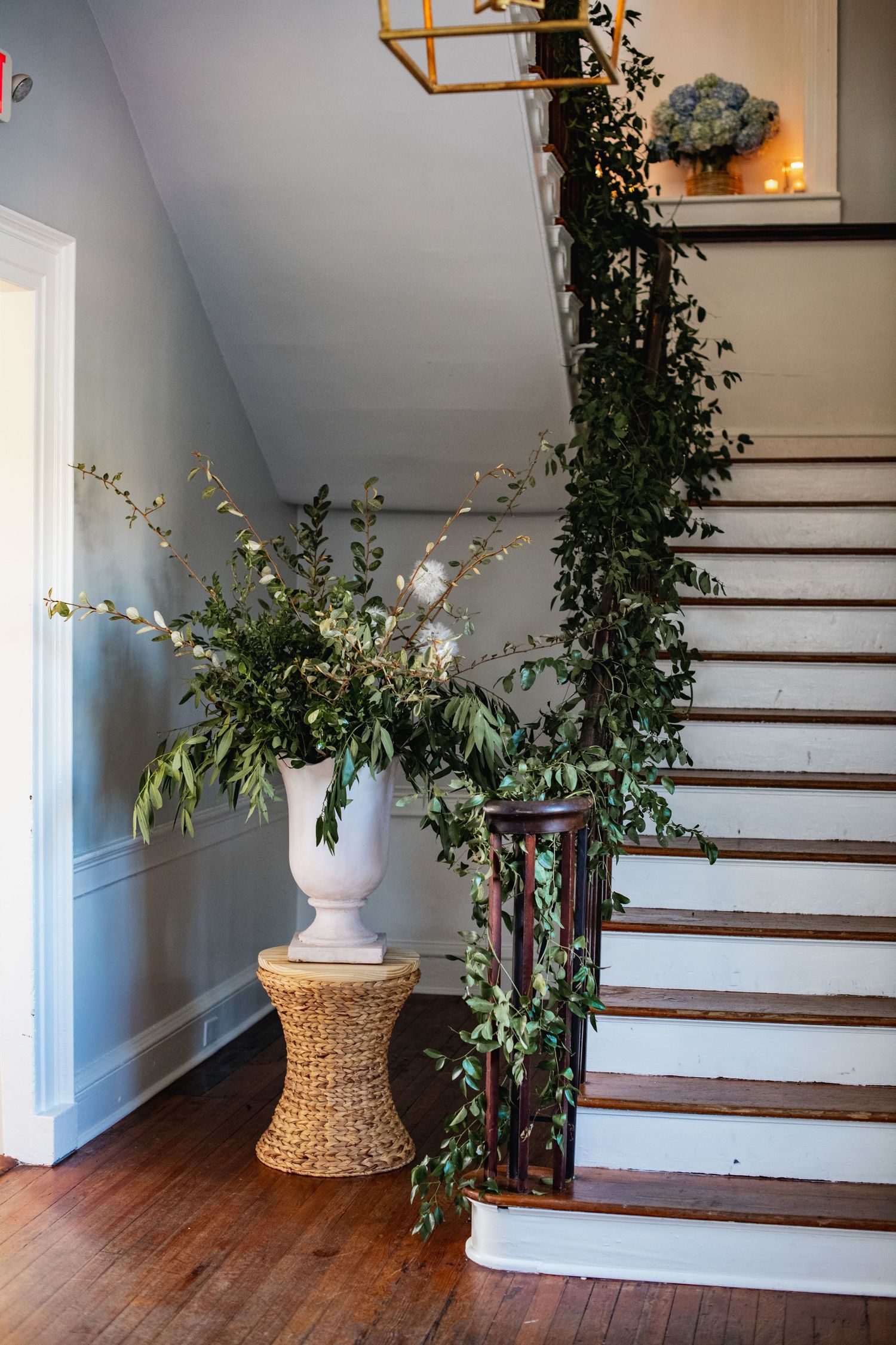A white vase with fresh greenery sits on a woven rattan stool beside a wooden staircase with climbing ivy.