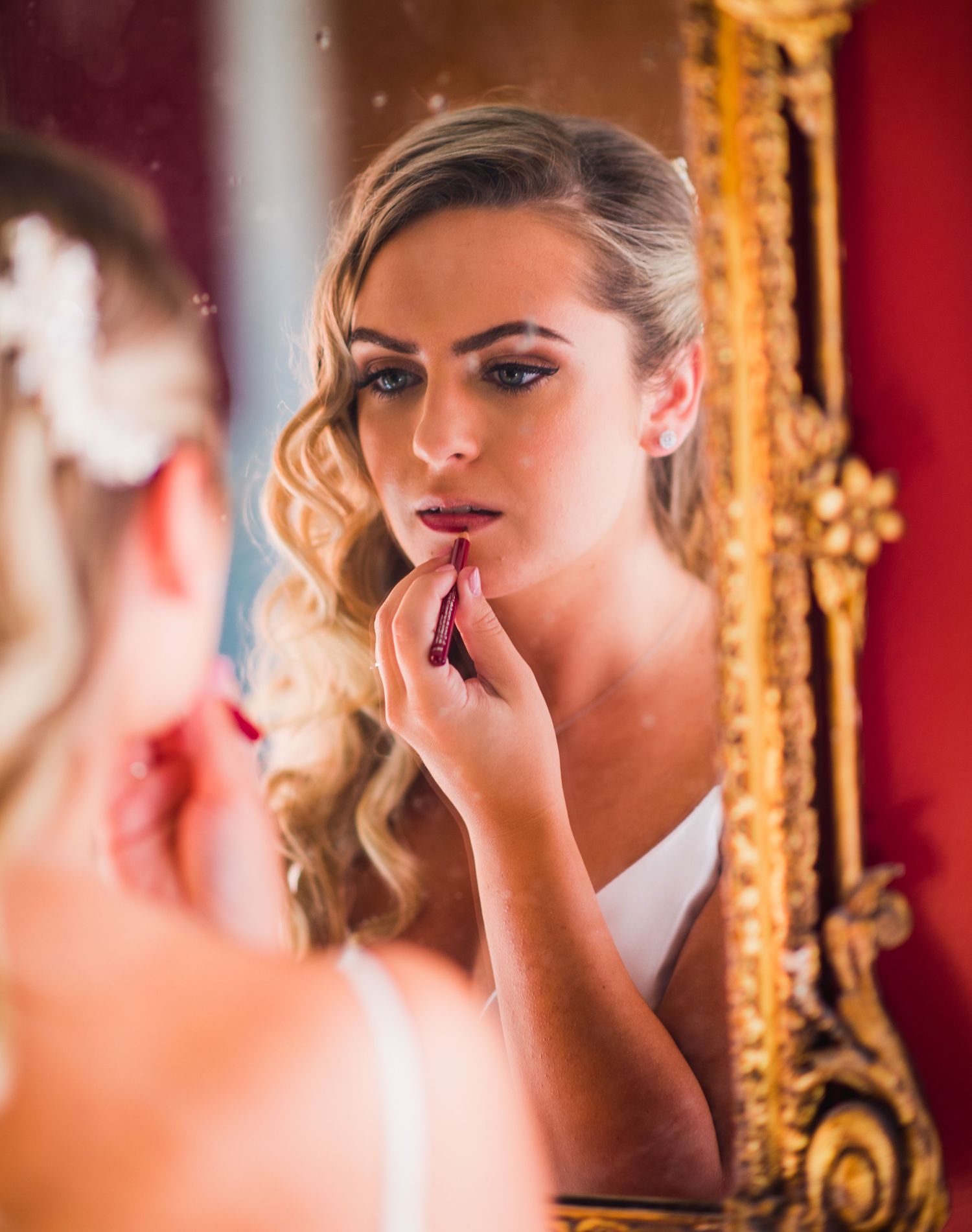 A person applies makeup while looking in an ornate gold-framed mirror against a red background.
