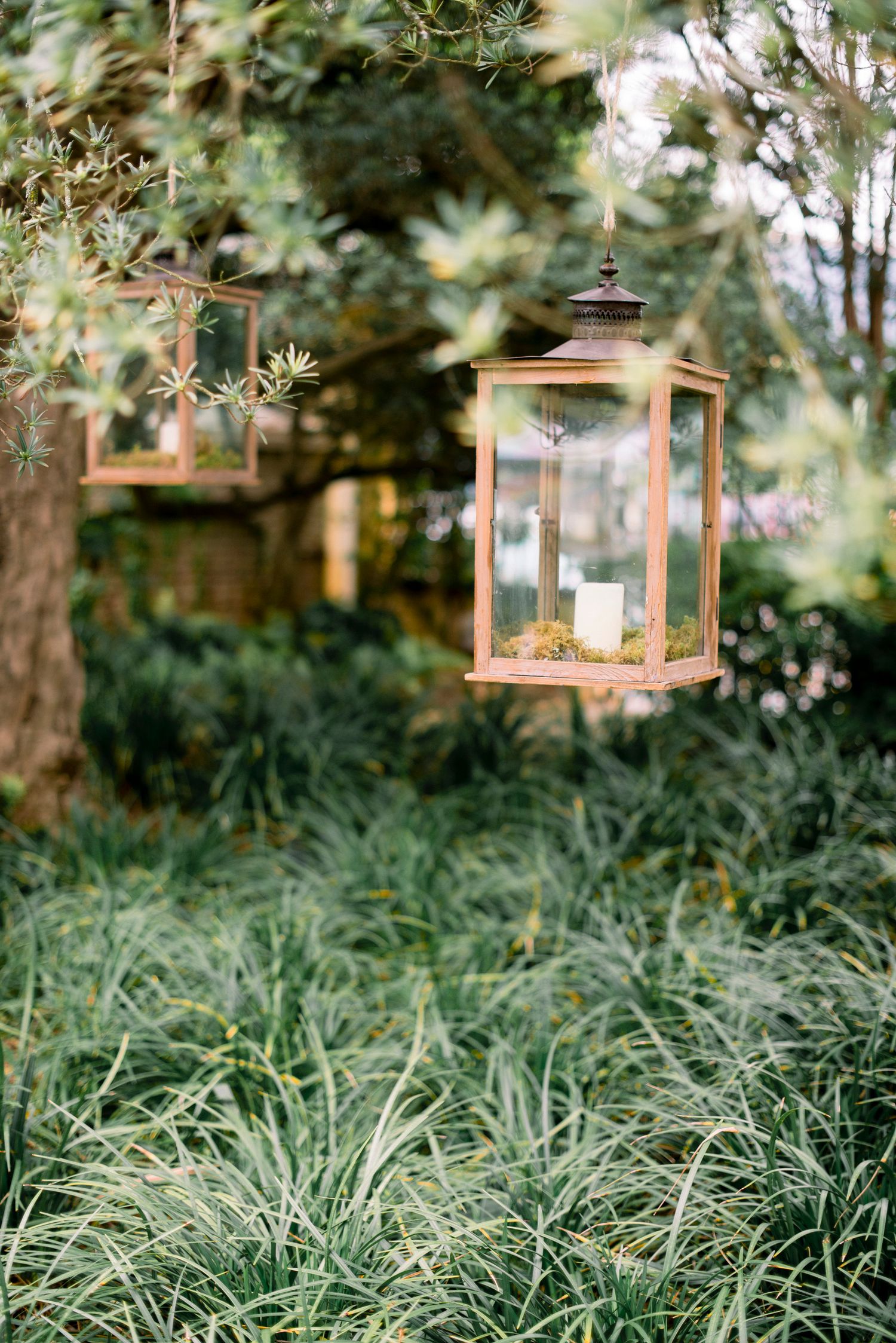 Decorative copper lanterns hang from poles in a lush garden setting with tall grass and trees.