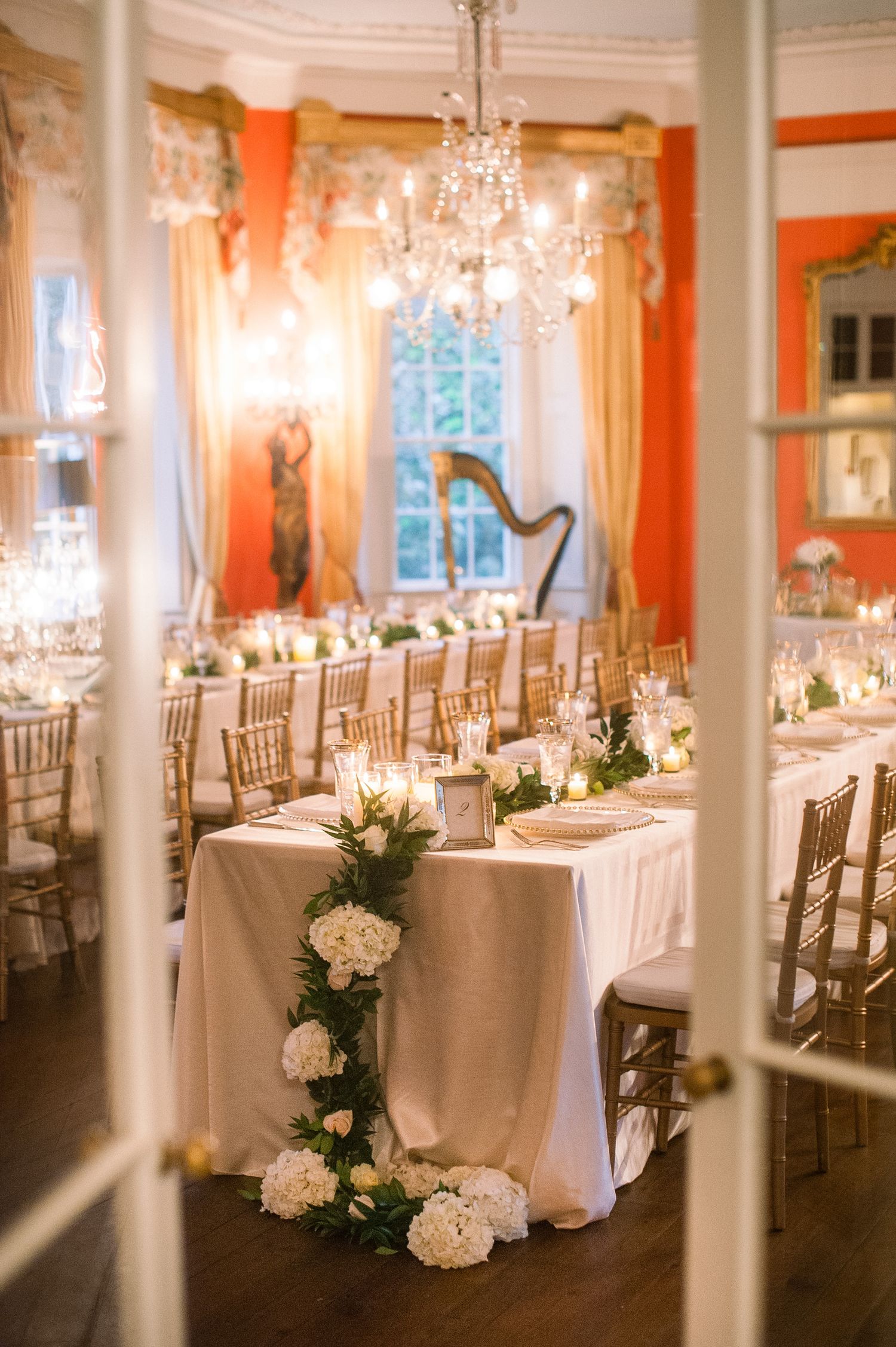 Sunlit banquet hall with coral-colored walls features rows of white-clothed tables and gold chiavari chairs for an elegant event.