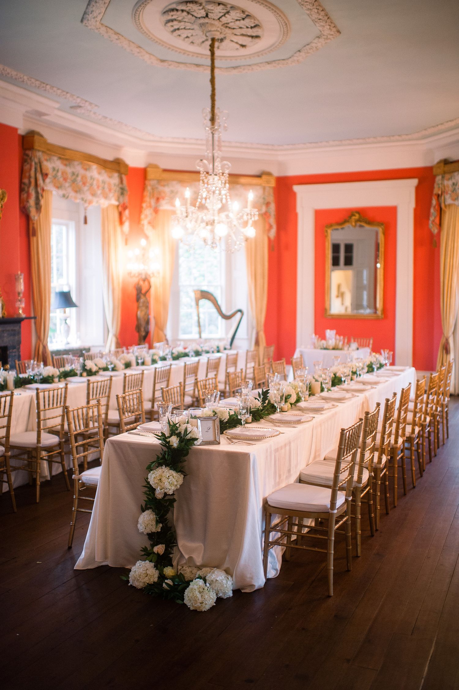 Elegant wedding reception room with coral walls, crystal chandeliers, and long tables set with white linens and gold chairs.