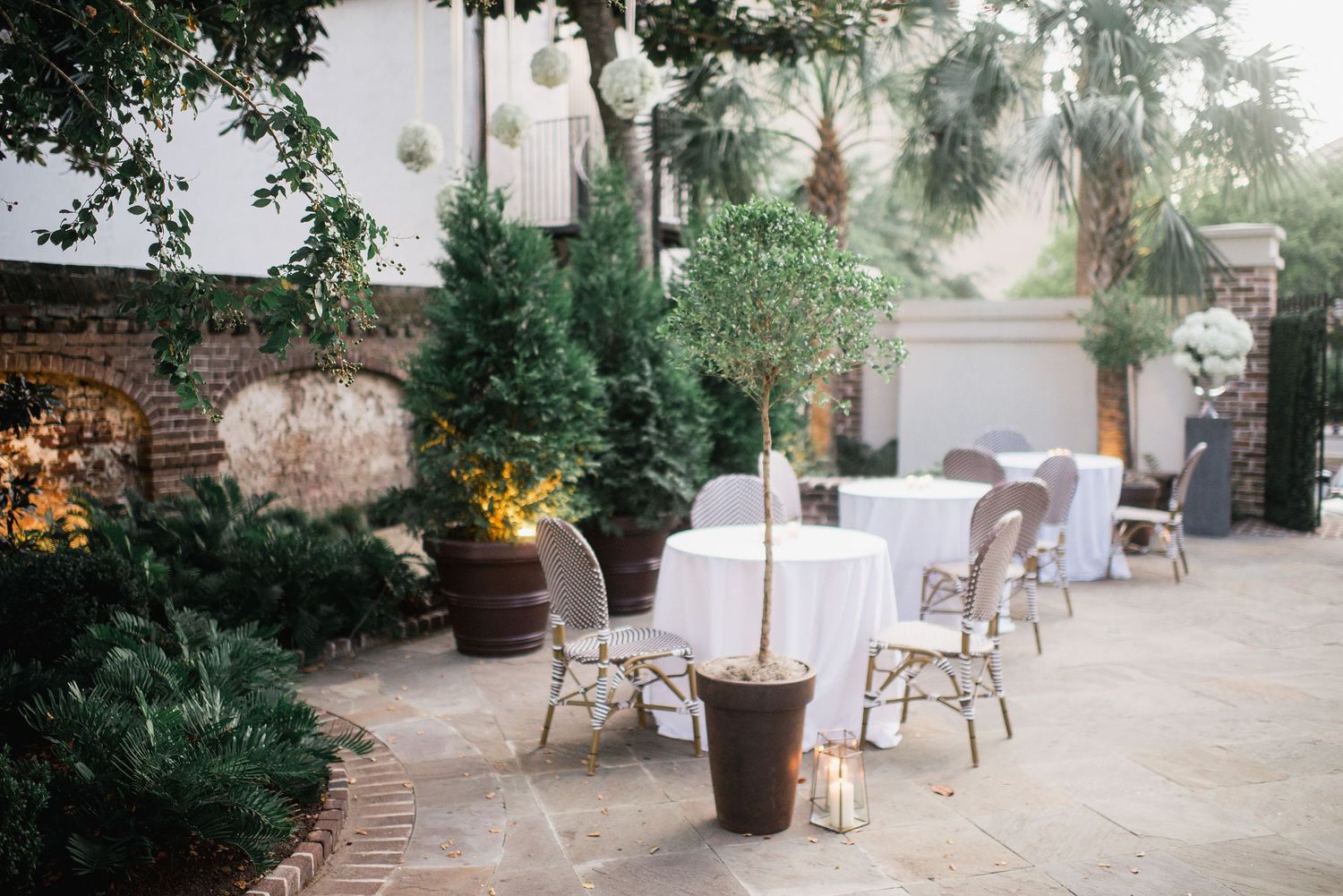 Elegant outdoor patio with white chairs and potted plants illuminated by warm lantern lighting in a garden setting.