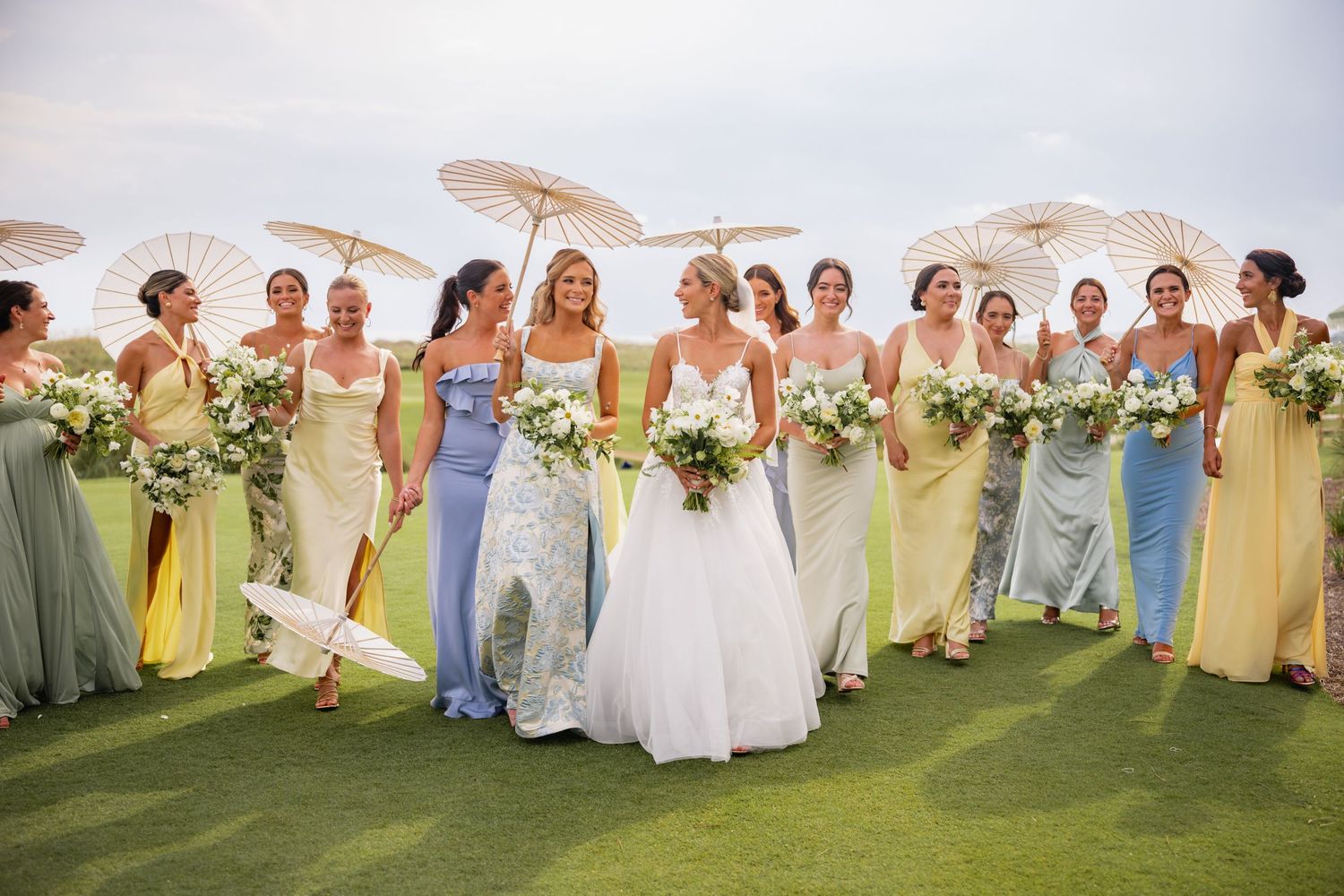 Wedding party in yellow and white dresses pose together on a sunny outdoor lawn for group photos.