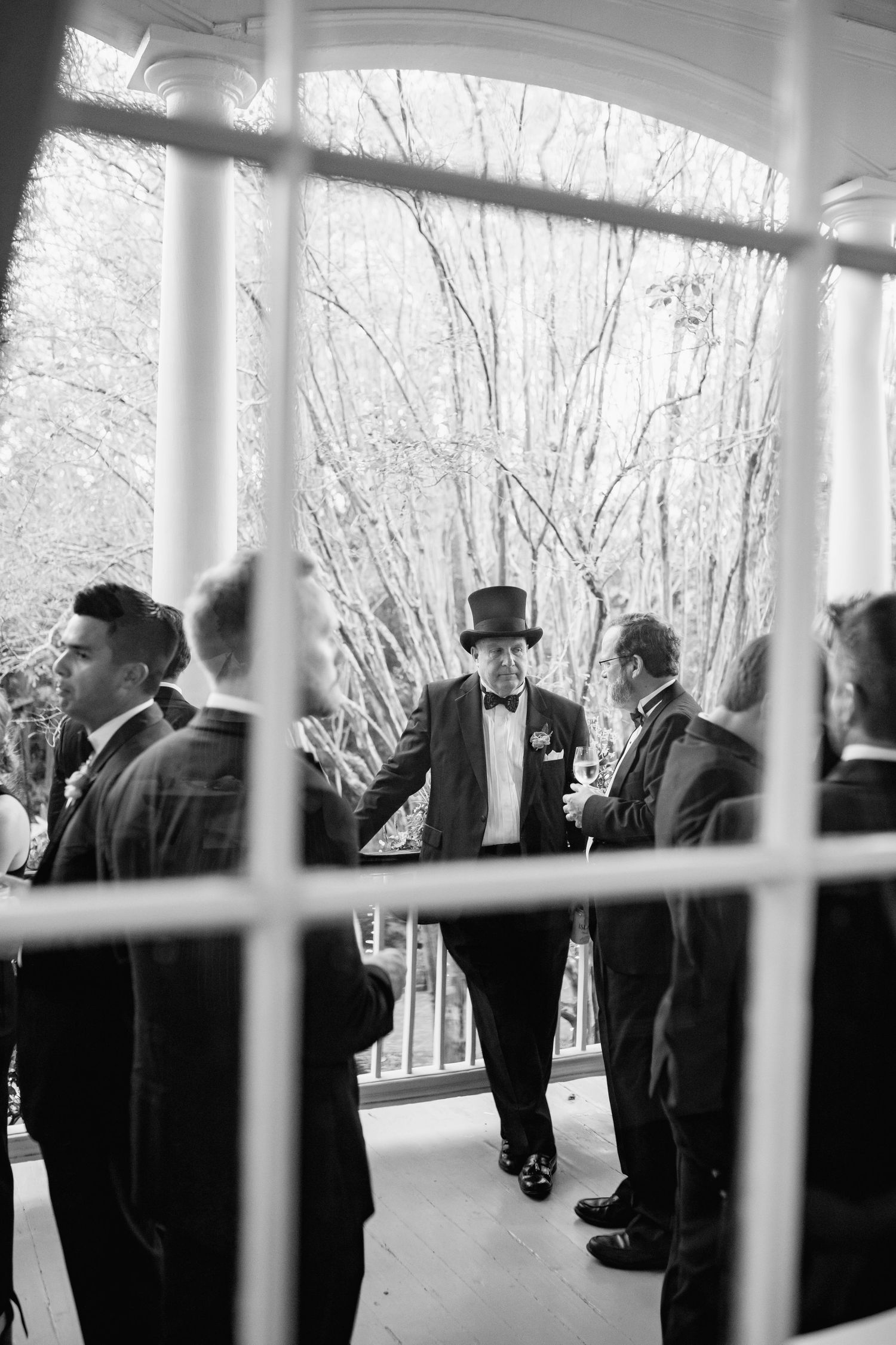 Black and white photo of people in formal attire and top hats gathering on a covered porch or veranda.
