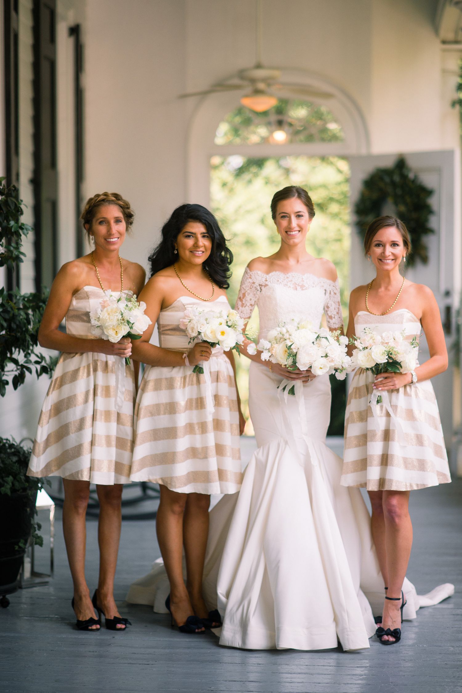 Bridesmaids in striped pink and white dresses pose with bouquets alongside bride in elegant white gown.