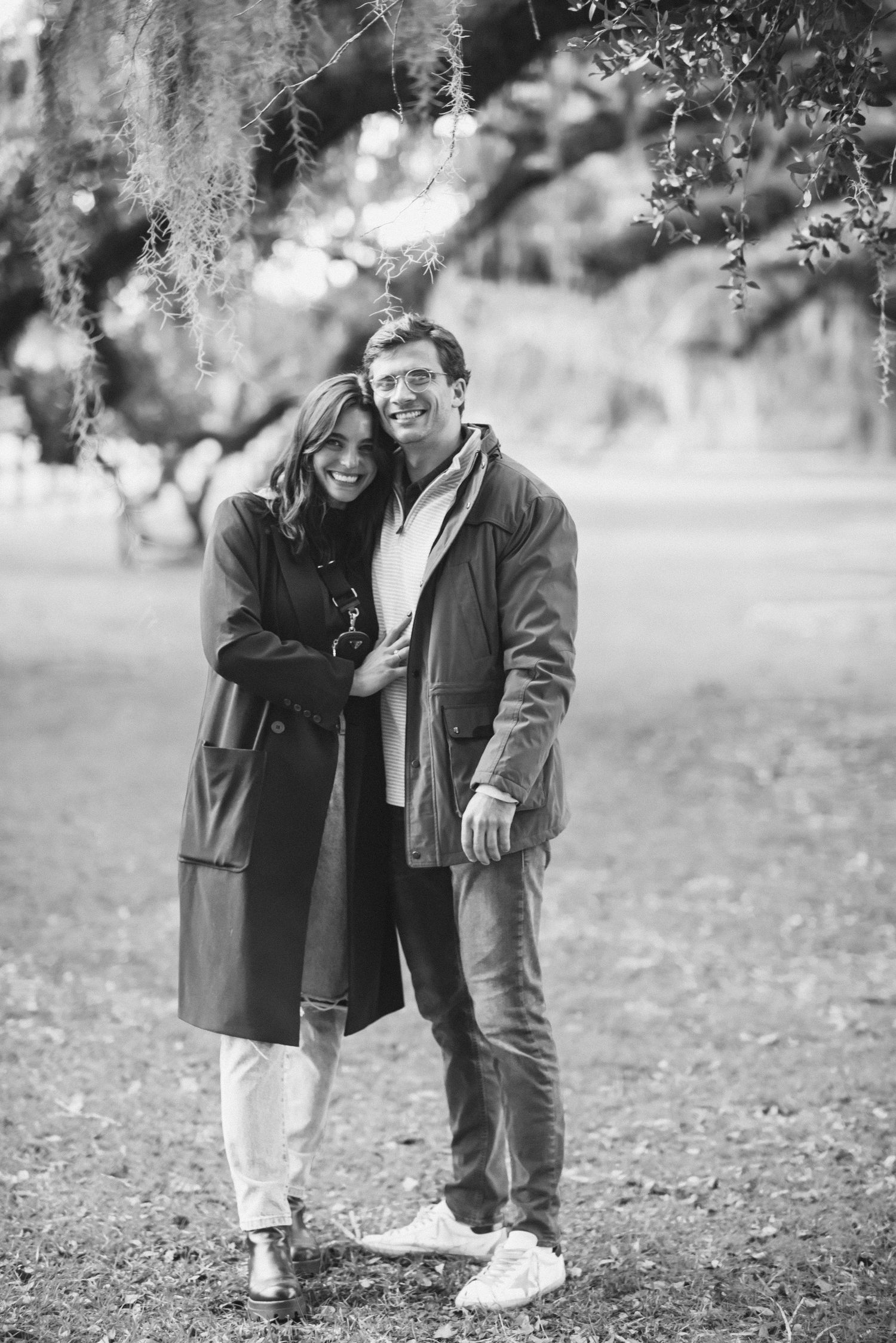 A couple shares a tender moment outdoors in black and white while standing together in a park setting.