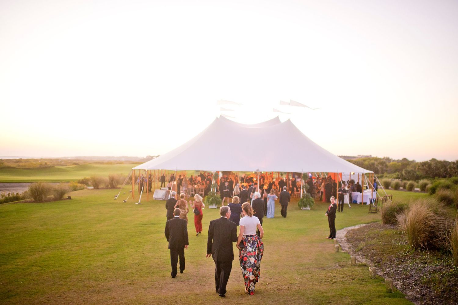 Guests walking towards a large white wedding tent set up on a scenic grassy hilltop during golden hour.