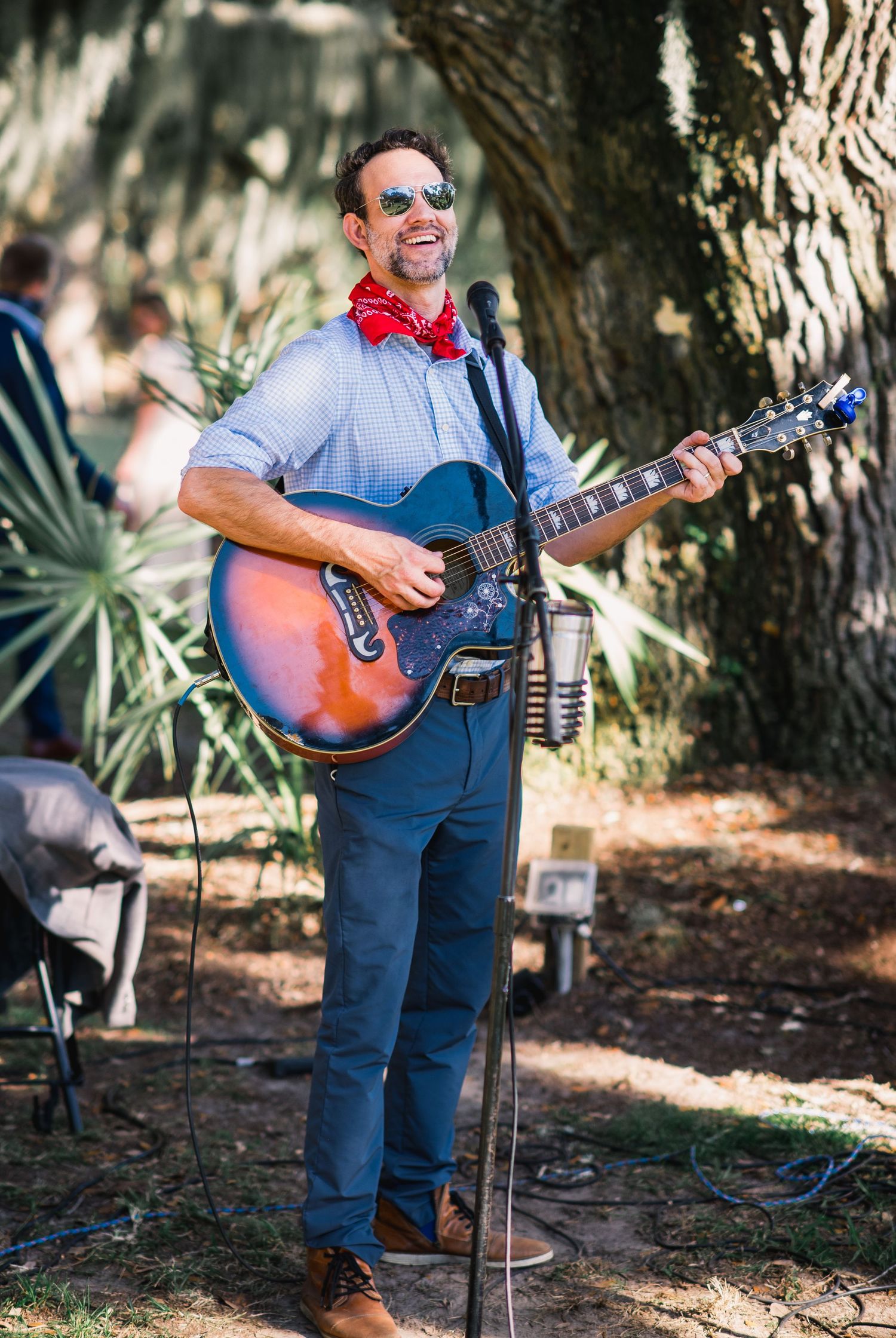 Musicians perform with acoustic instruments under shaded trees at an outdoor bluegrass festival.
