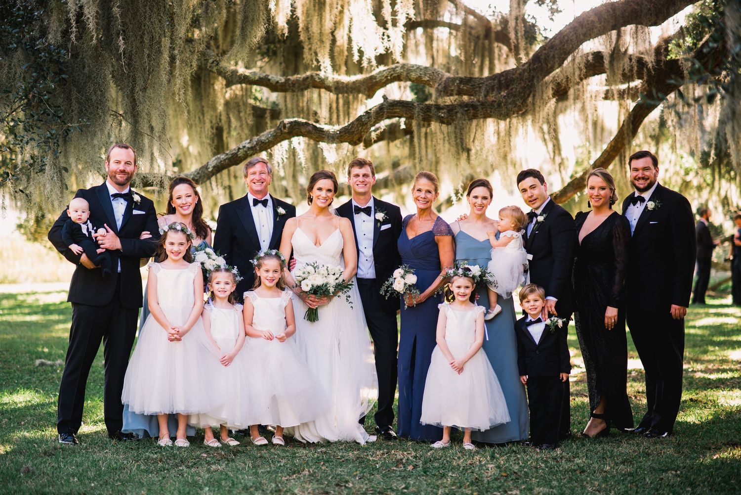 Wedding party poses together under moss-draped oak trees in a romantic garden setting.