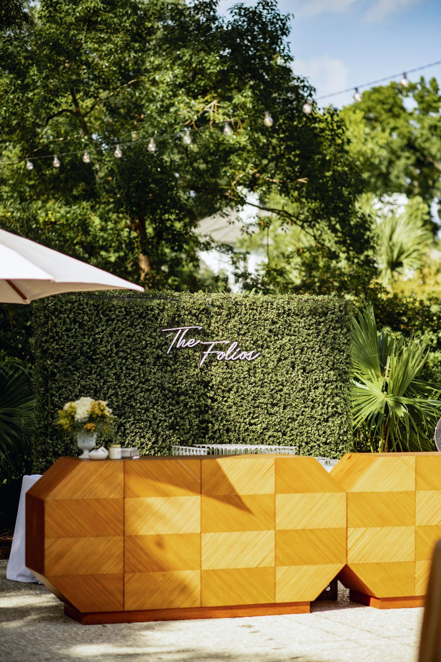 Modern yellow geometric bar setup with hedge wall backdrop and white umbrella in an outdoor garden setting.