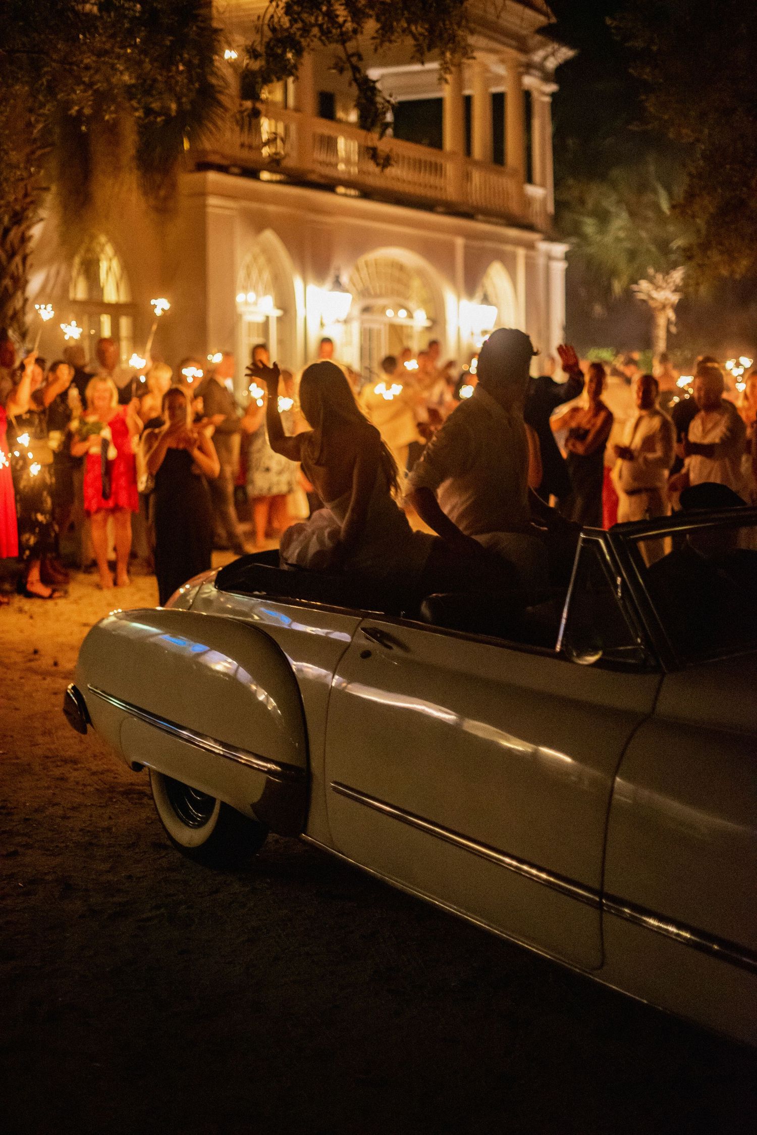 A vintage car parked at a nighttime wedding celebration with sparklers glowing as guests gather outside an elegant venue.