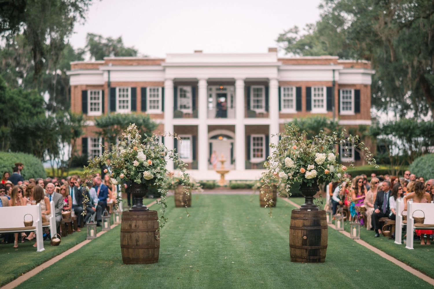 Wedding guests watch a ceremony on a sprawling green lawn in front of a historic colonial-style mansion.