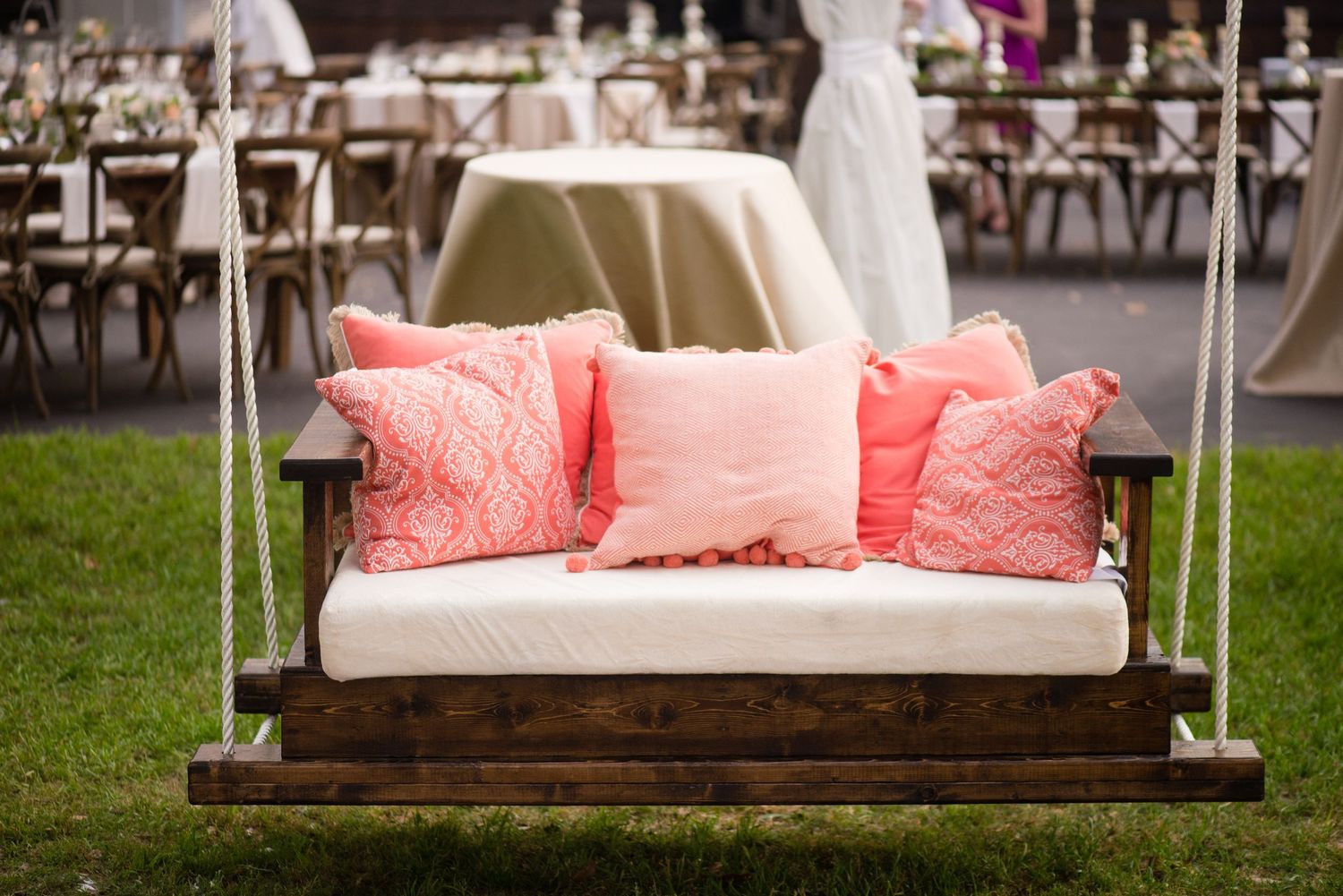A wooden porch swing with coral and white cushions hangs by rope against a garden backdrop.
