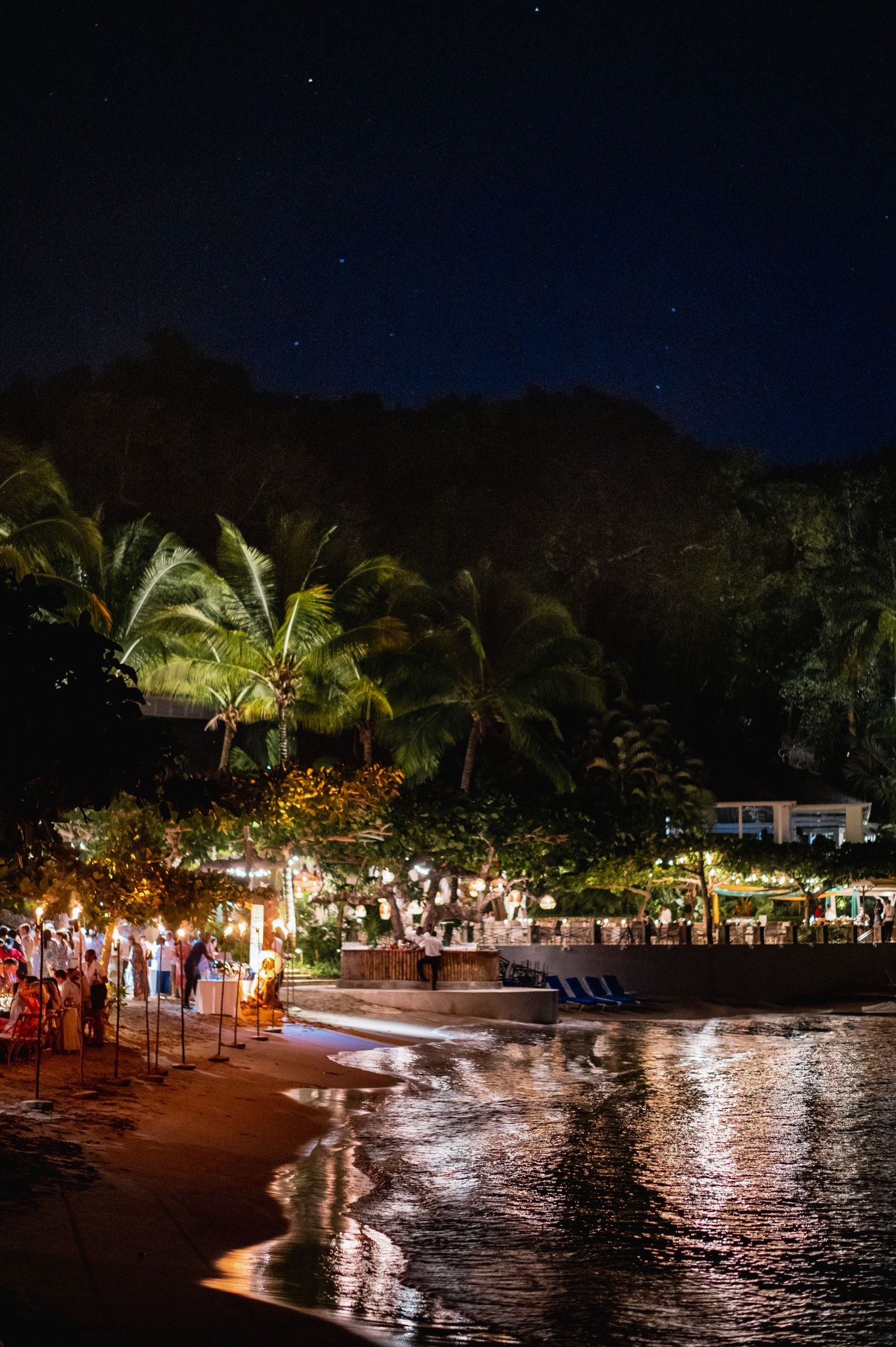 Nighttime panorama of a tropical beach resort with illuminated palm trees and reflections on calm water.