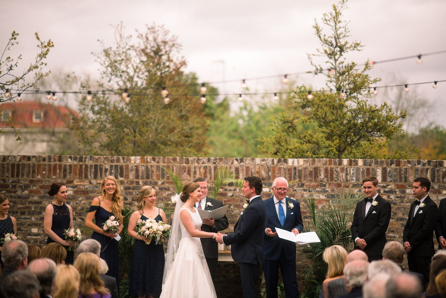 An outdoor wedding ceremony takes place at dusk with string lights and trees creating a romantic garden atmosphere.