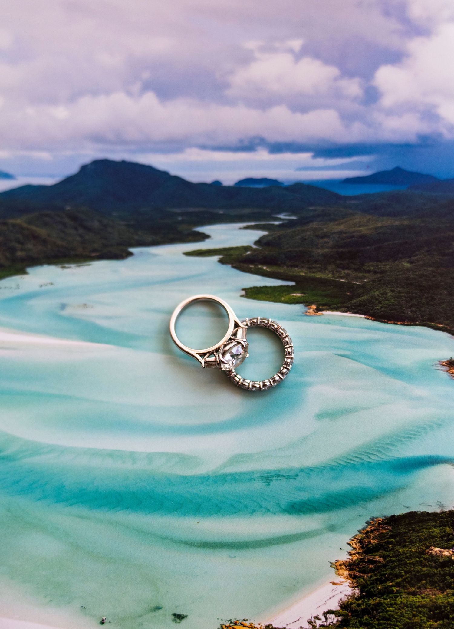 Wedding rings displayed against a stunning aerial view of turquoise waters and mountains in a tropical landscape.