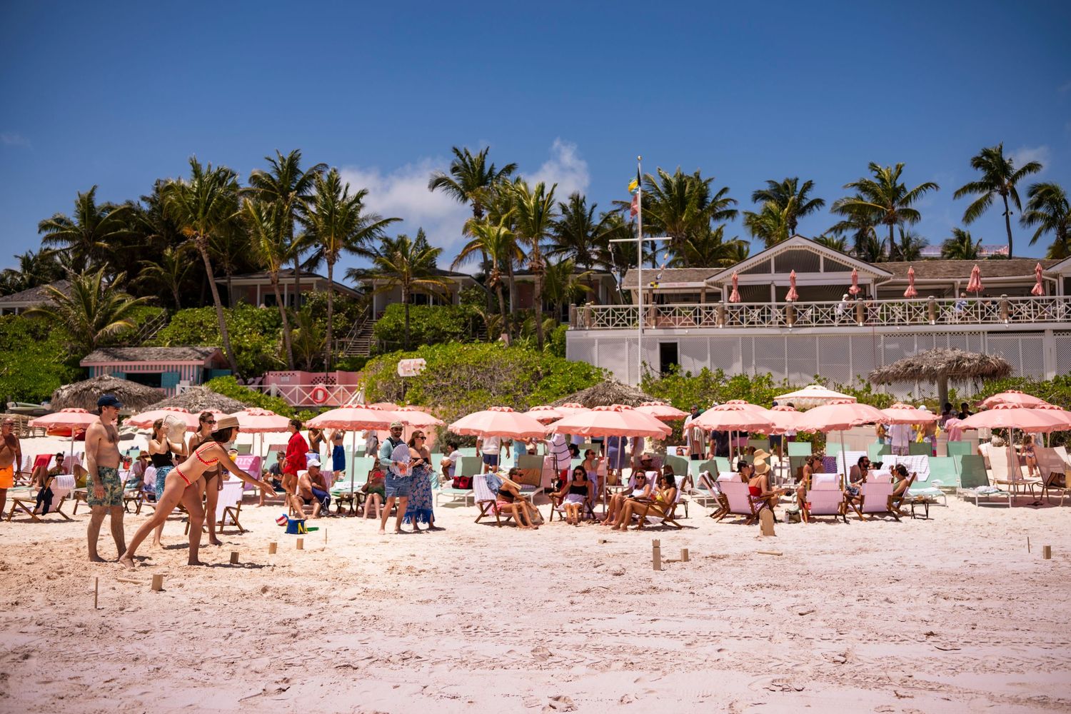Crowded beach scene with pink umbrellas and palm trees at a tropical resort.