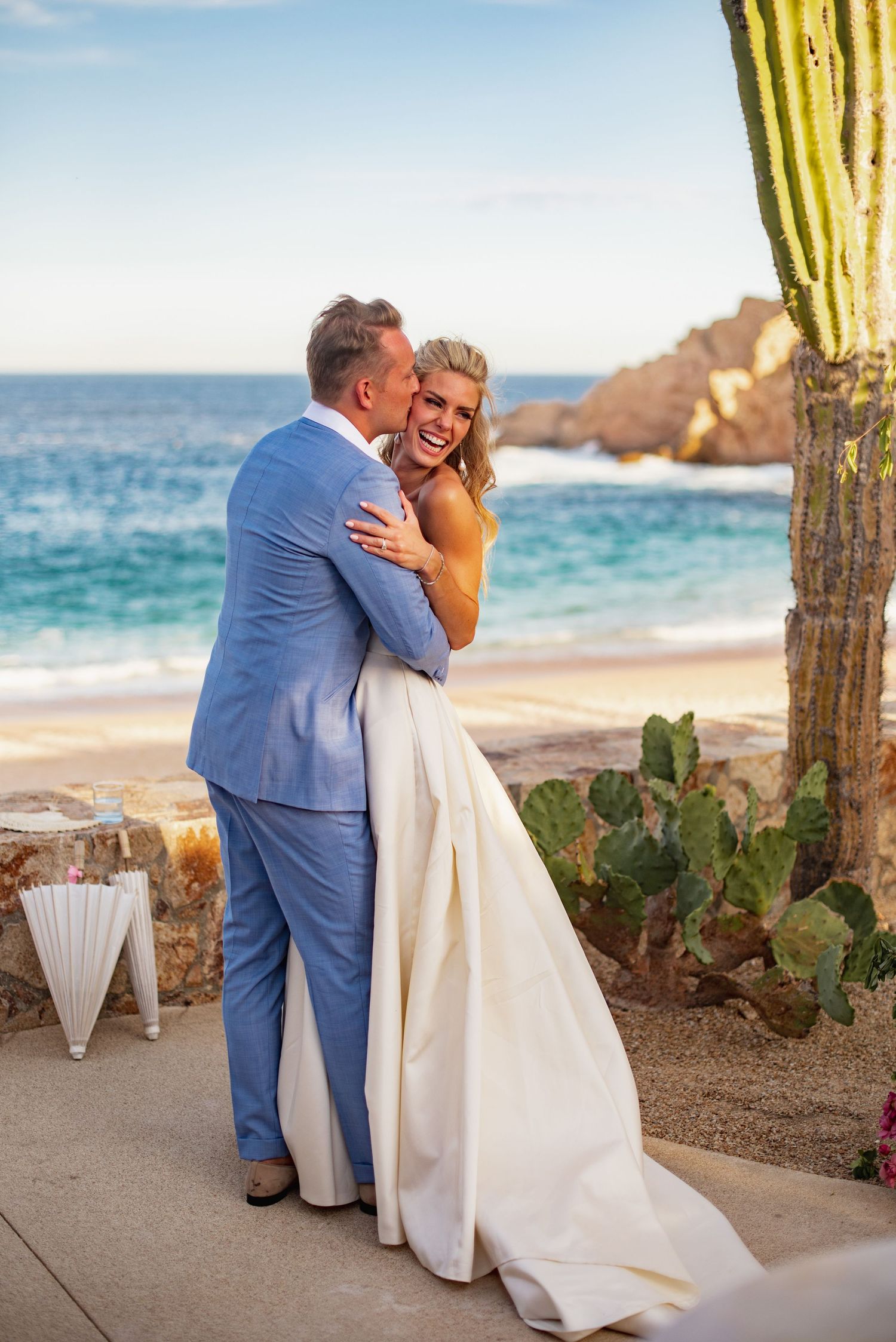 A couple embraces on a beach at sunset with cactus plants and ocean views in the background.