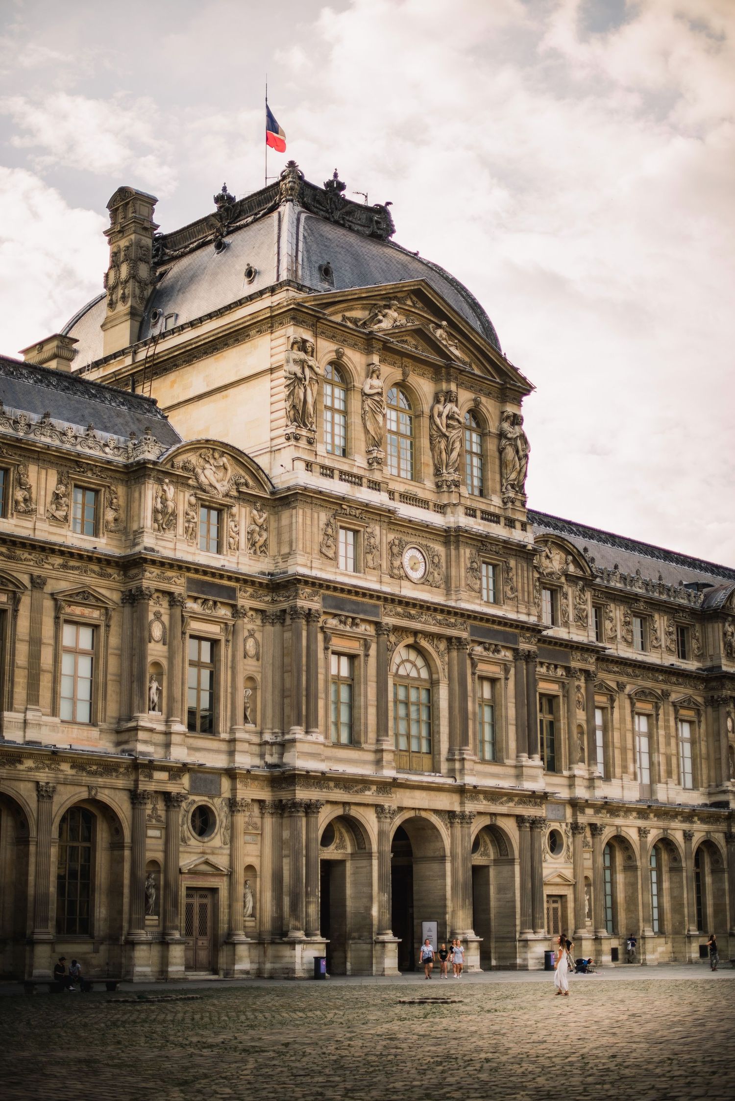 The ornate facade of the Louvre Museum in Paris with its classical French architecture and tricolor flag flying above.
