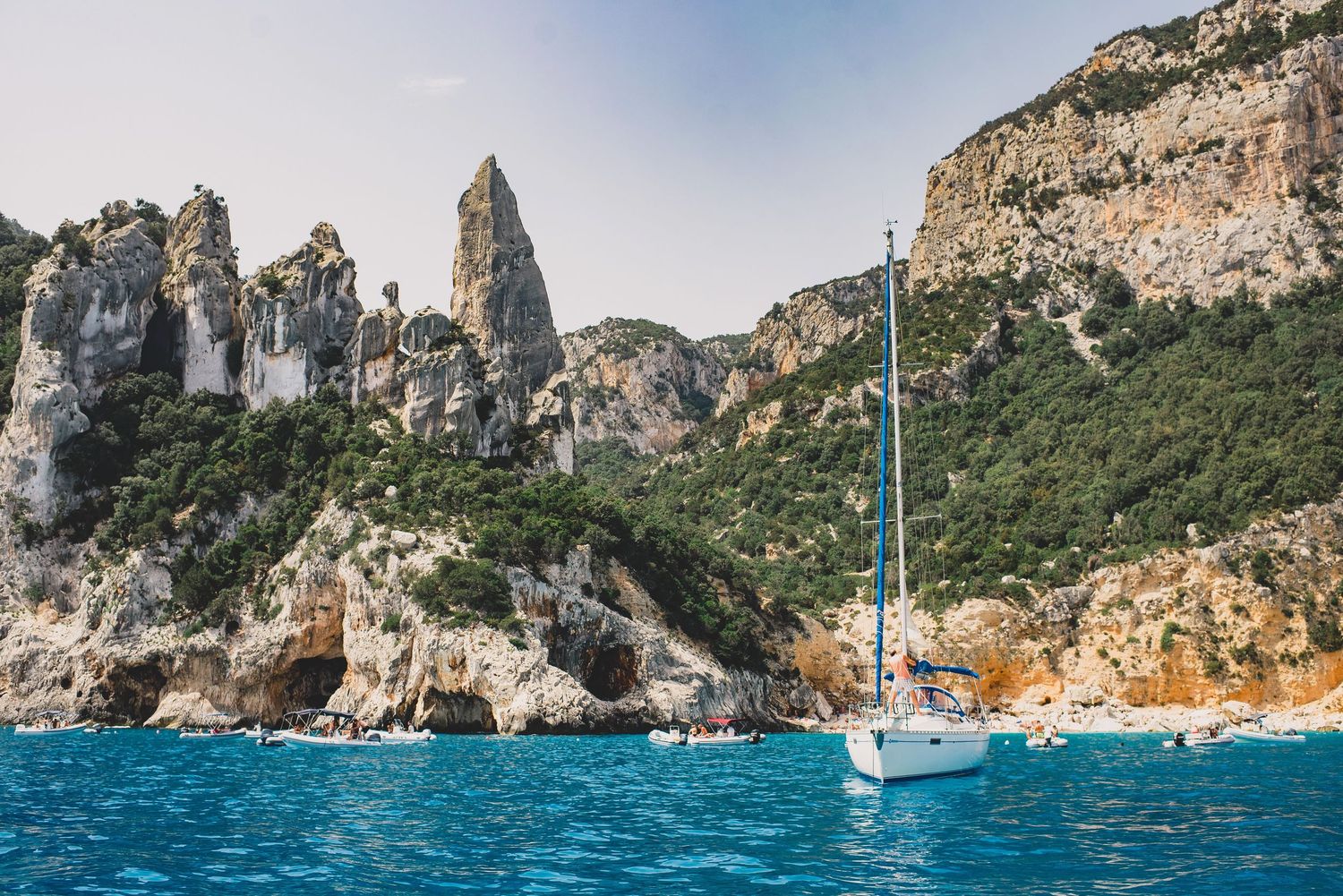 Rocky coastal cliffs and turquoise waters of the Mediterranean Sea with sailboats anchored near the shore.