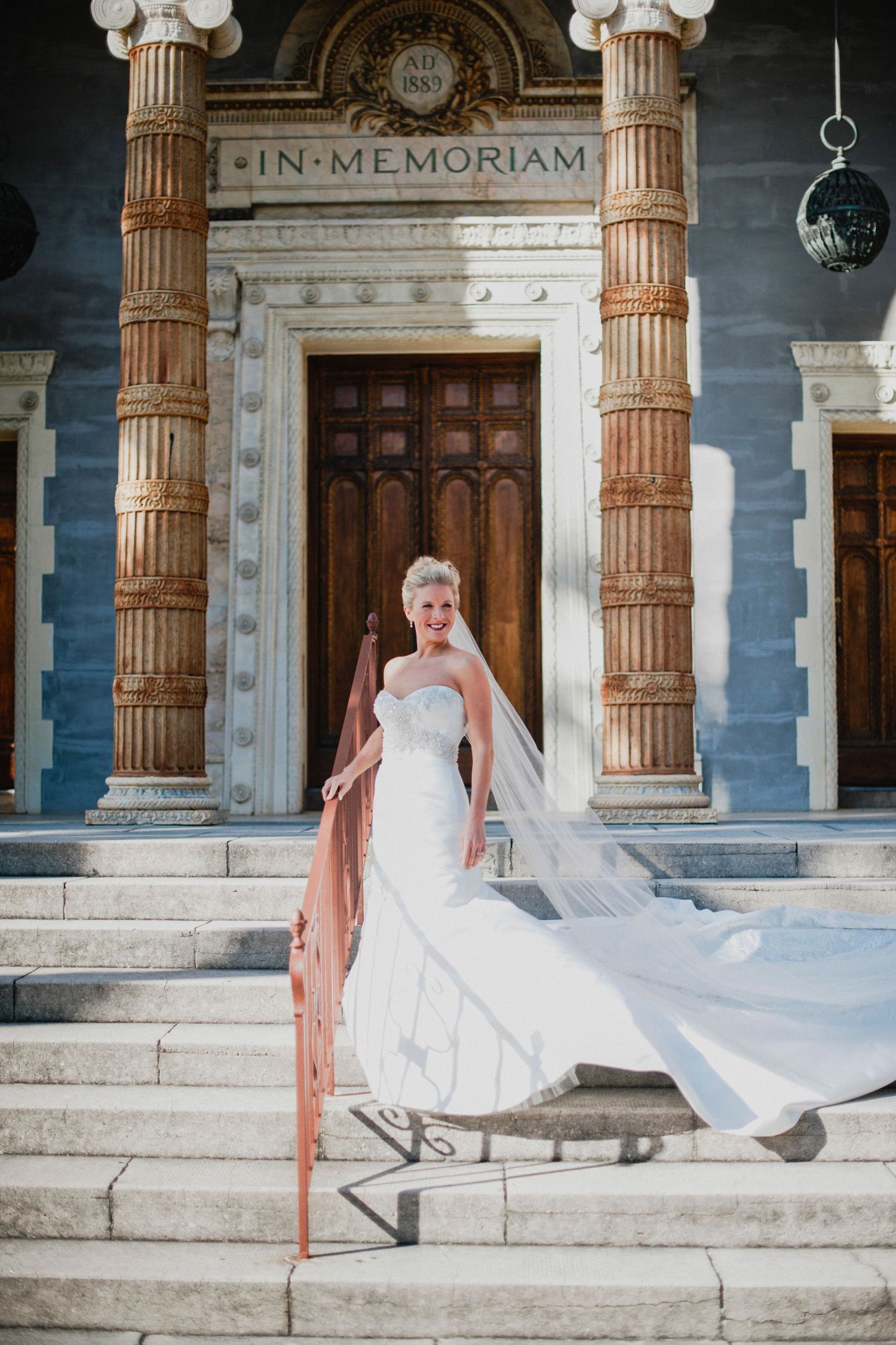 A bride in a flowing white dress poses gracefully on marble steps of a classical building with ornate columns.
