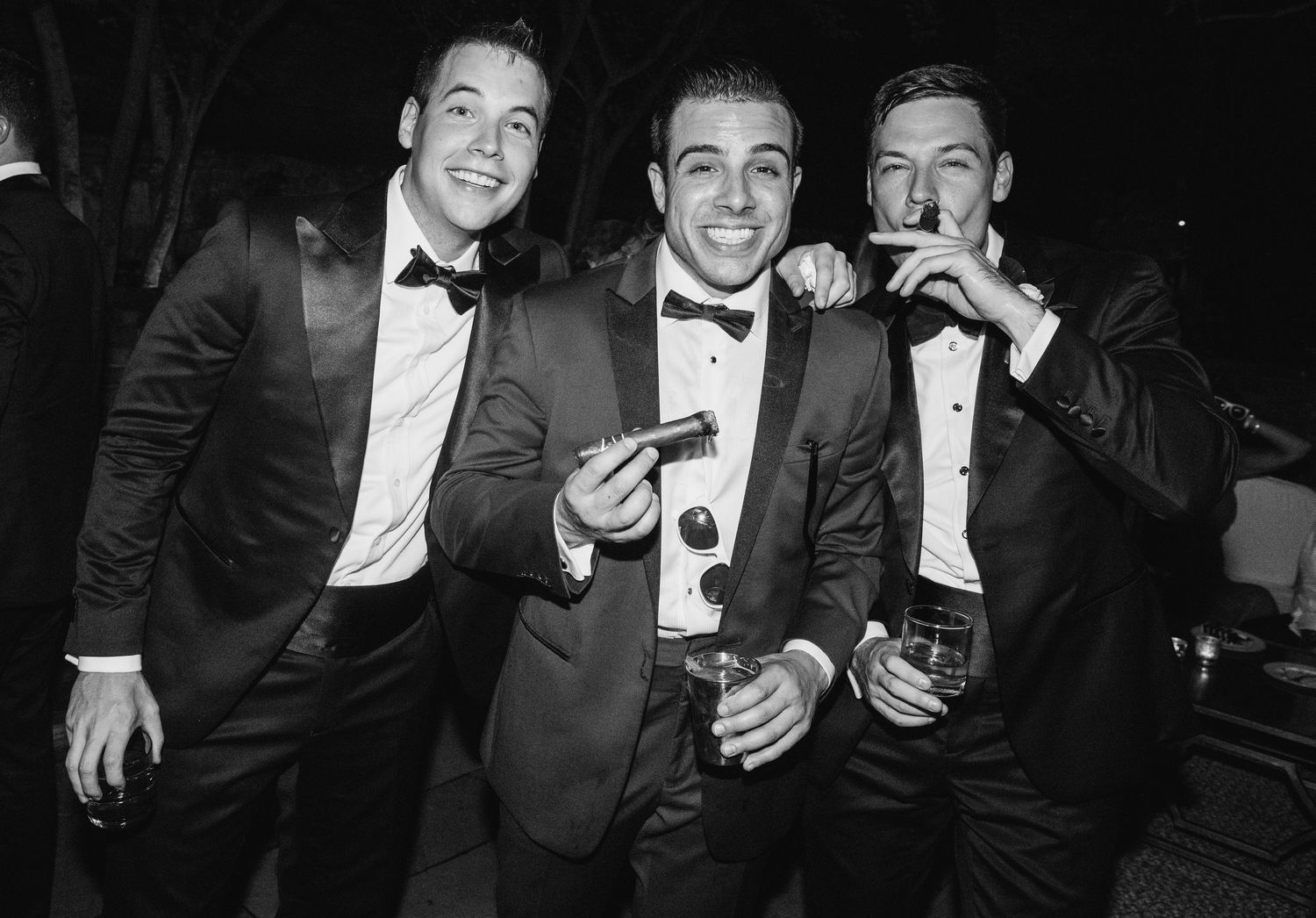 A group in formal black tie attire laughs and socializes at an elegant evening event.
