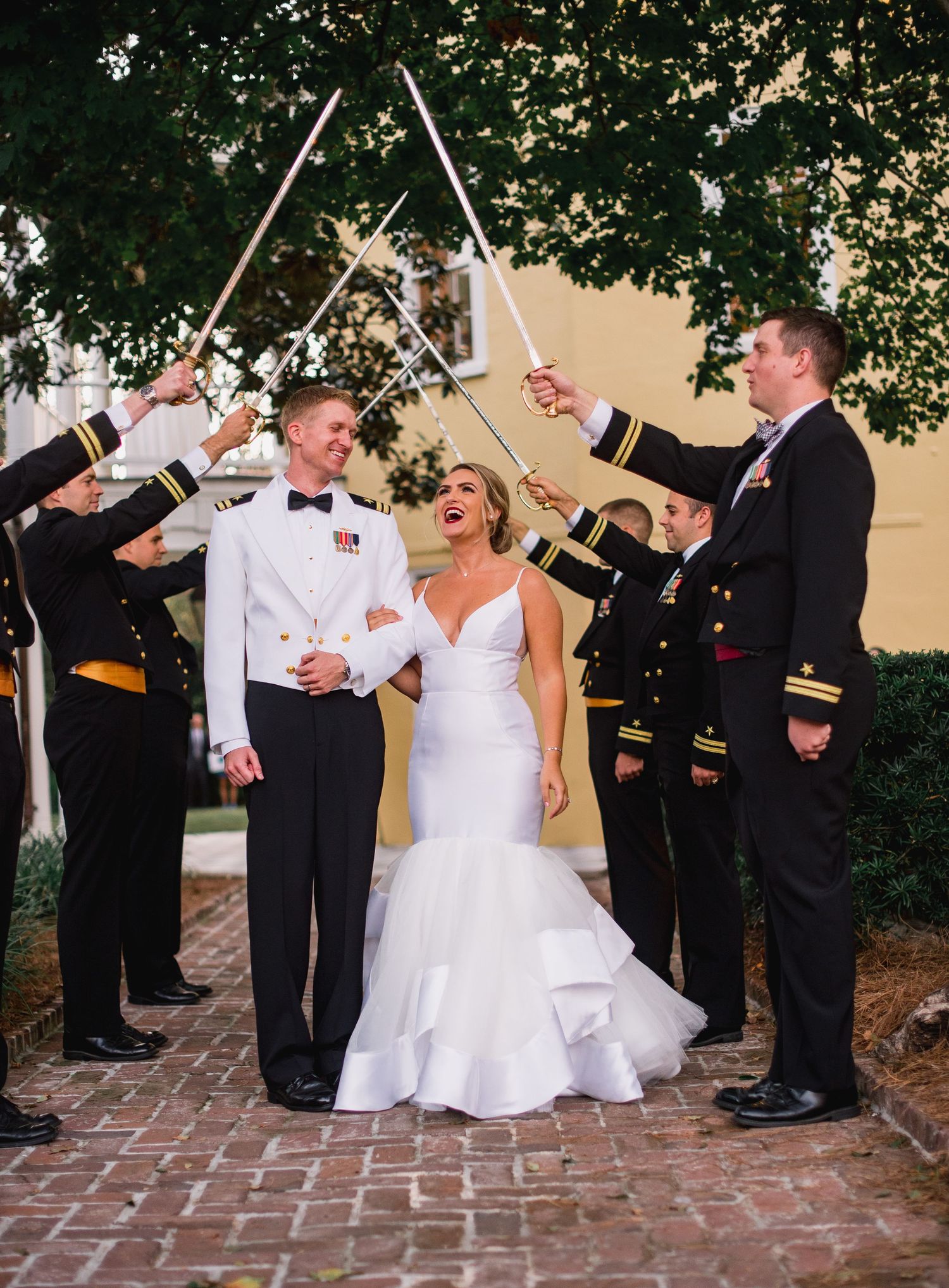 A wedding party line performs a fun synchronized drinking pose with bottles on a brick walkway outdoors.
