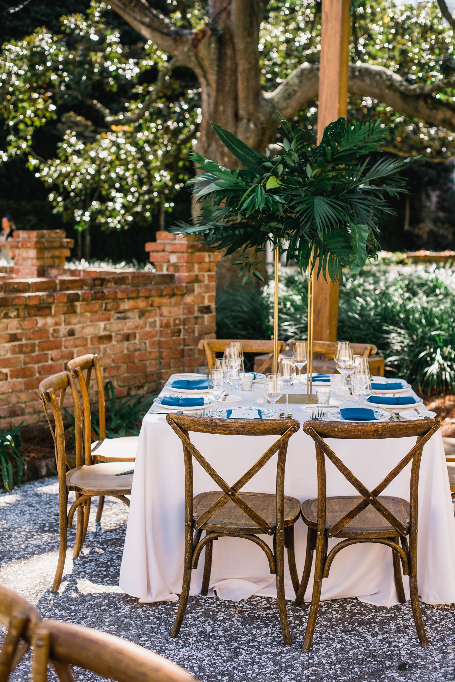 Elegant outdoor dining setup with wooden cross-back chairs and white tablecloths under flowering trees in a garden setting.