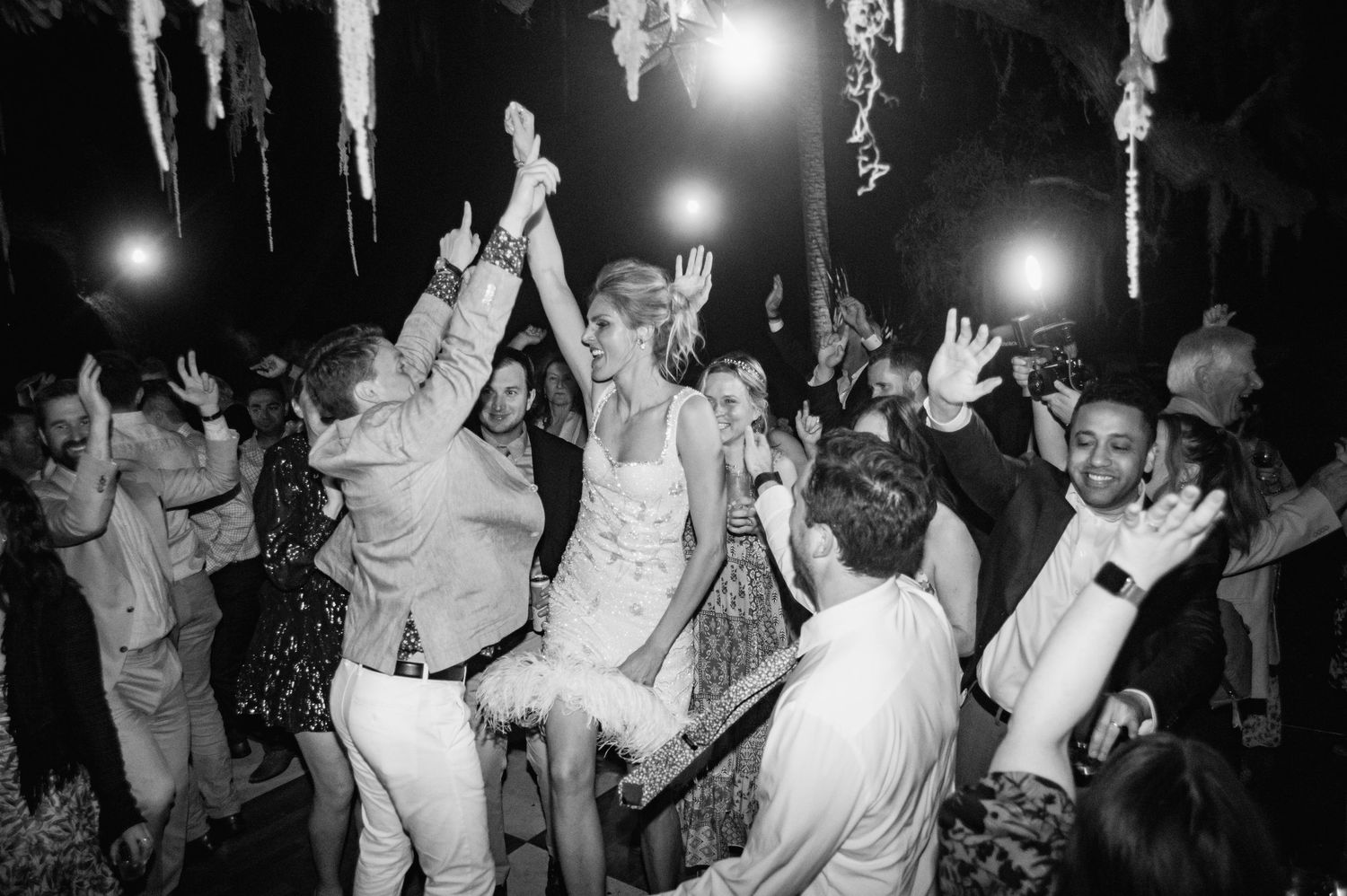 A black and white photo of people dancing and celebrating with their arms raised at a nightclub or party.