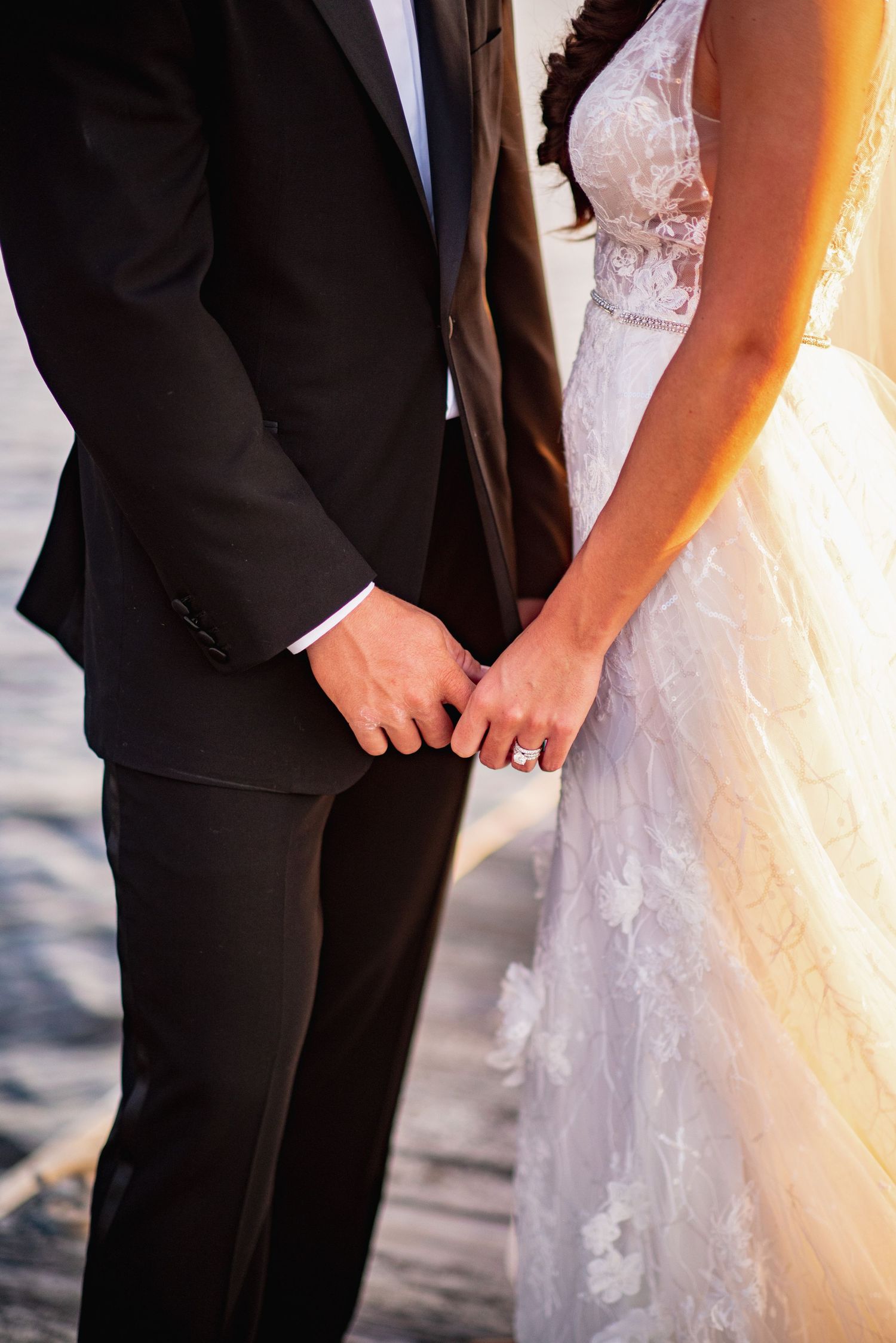 A bride and groom stand close together on a beach at sunset, with their formal wedding attire silhouetted against the light.