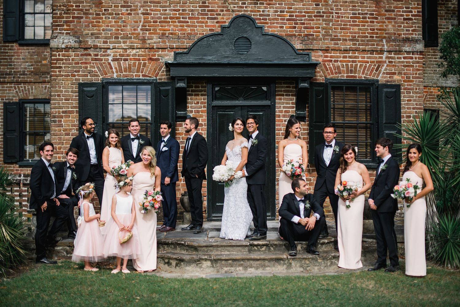 Wedding party poses in front of historic brick building with elegant formal attire and floral bouquets.