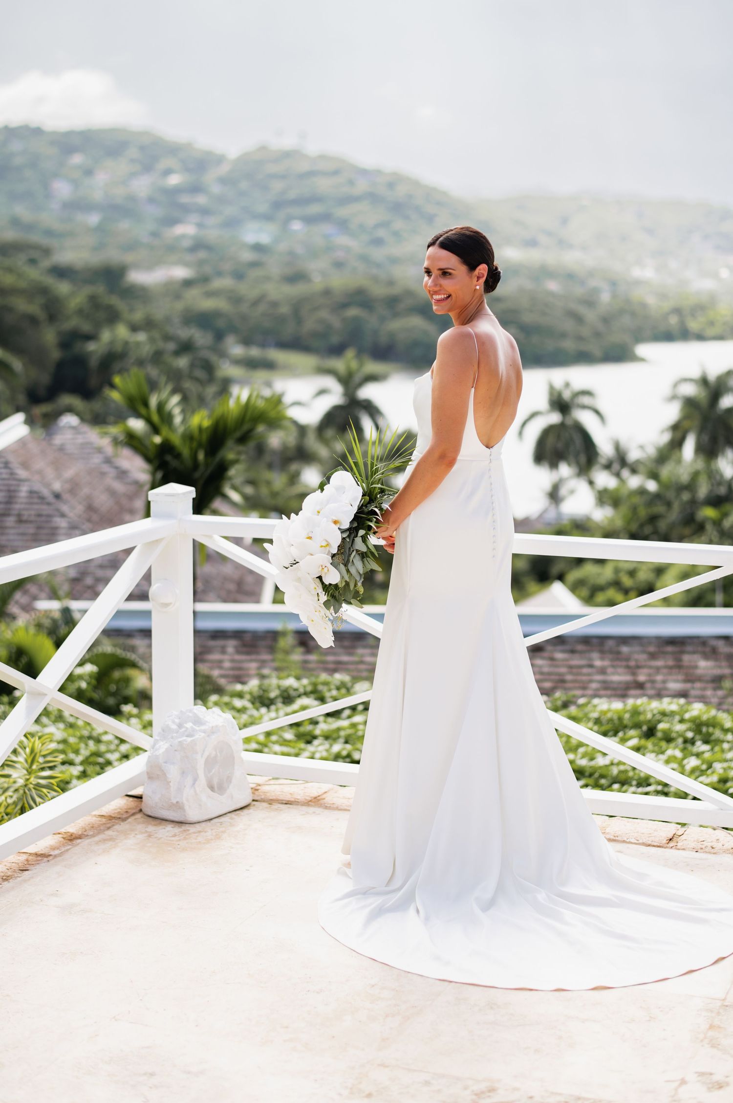 Elegant bride in sleek white backless wedding dress poses on tropical balcony overlooking ocean and palm trees.