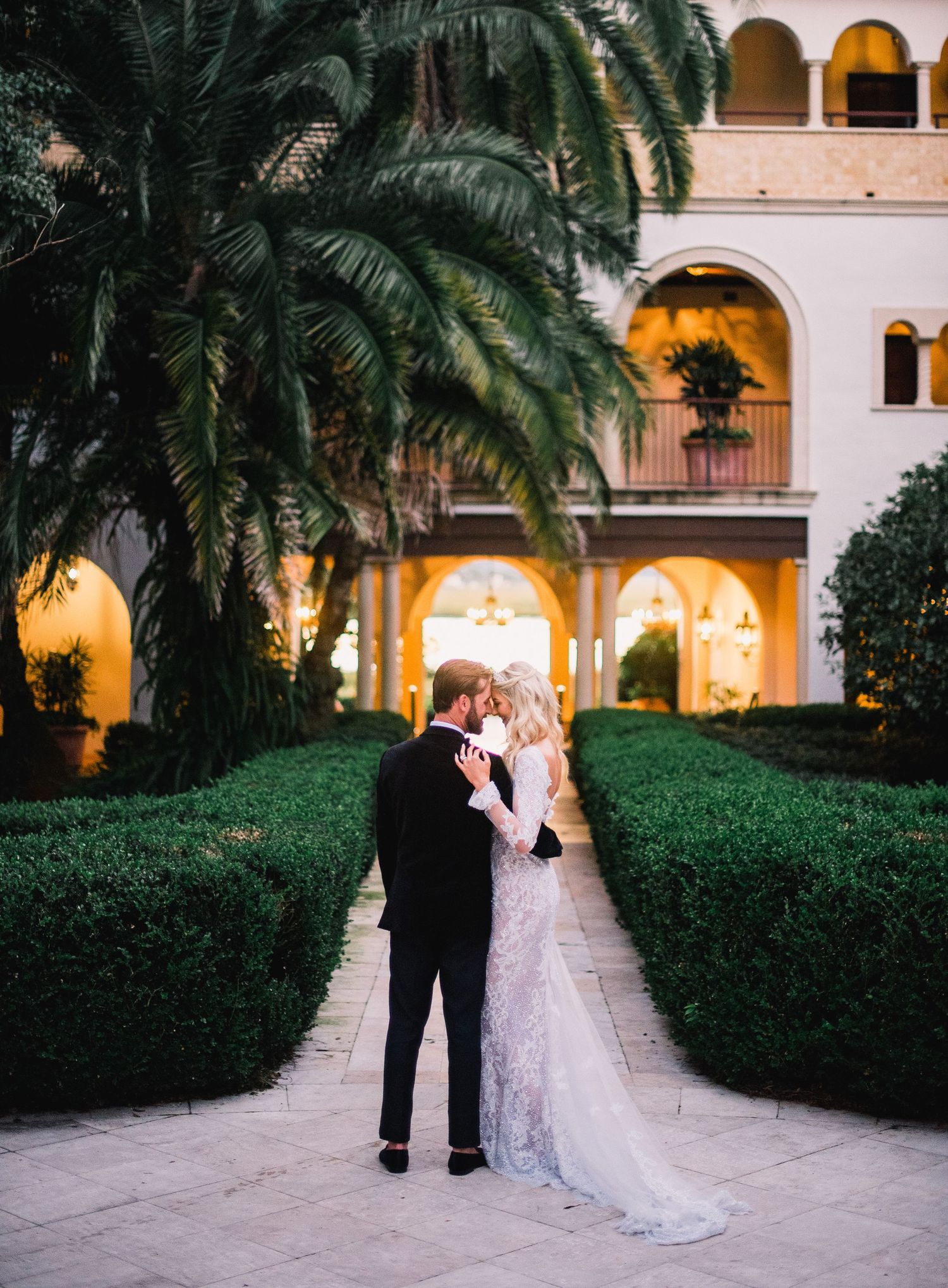 A couple shares an intimate moment in a romantic garden courtyard with palm trees and illuminated arches at dusk.