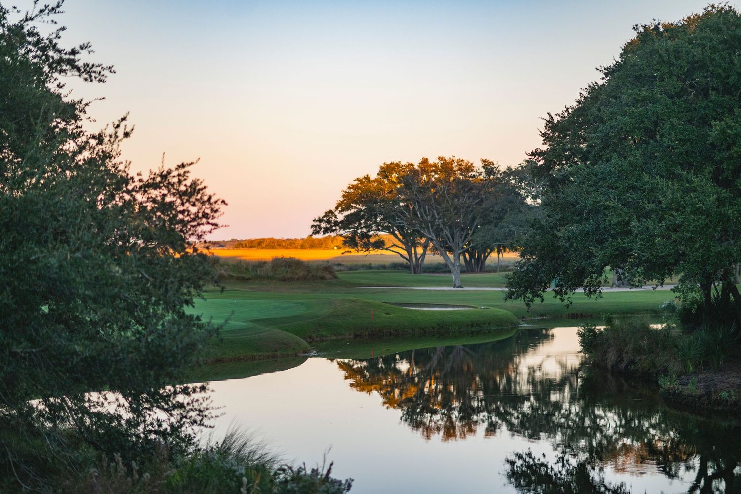 Scenic golf course landscape with water hazard and trees silhouetted against a colorful sunset sky.