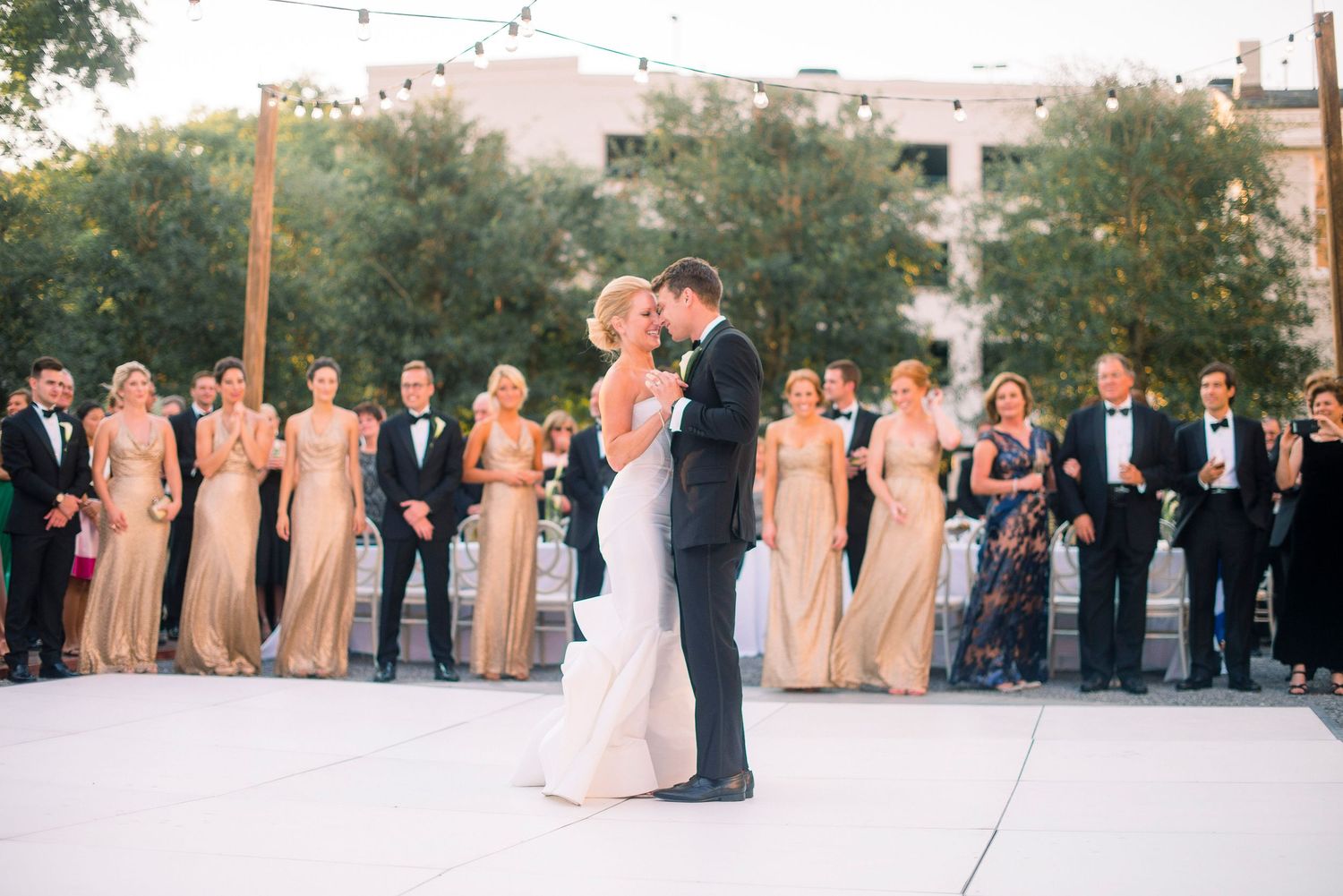 A couple shares their first dance outdoors at dusk surrounded by their wedding party in formal attire.