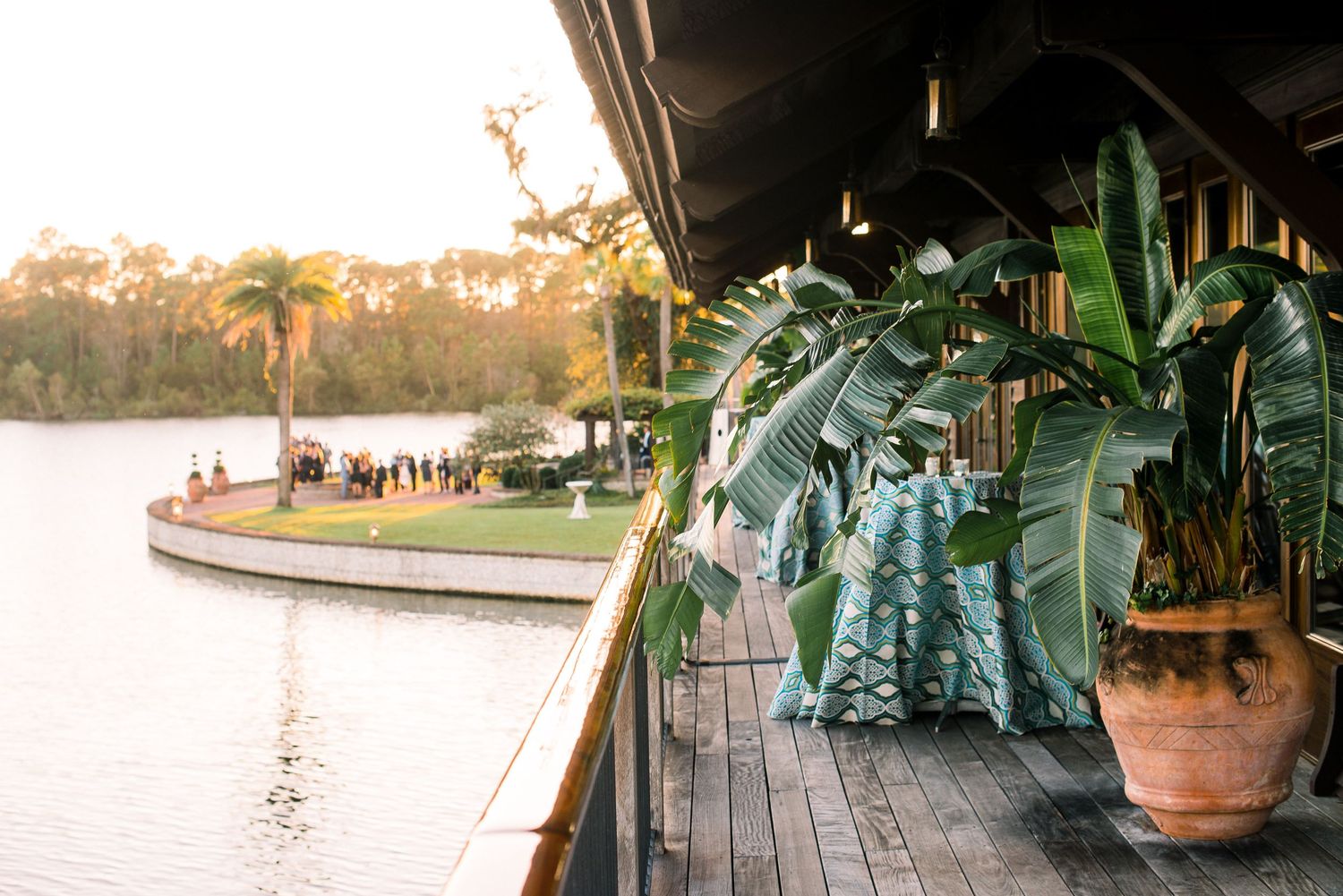 A serene wooden deck overlooking a tranquil lake at sunset with potted tropical plants and palm trees.