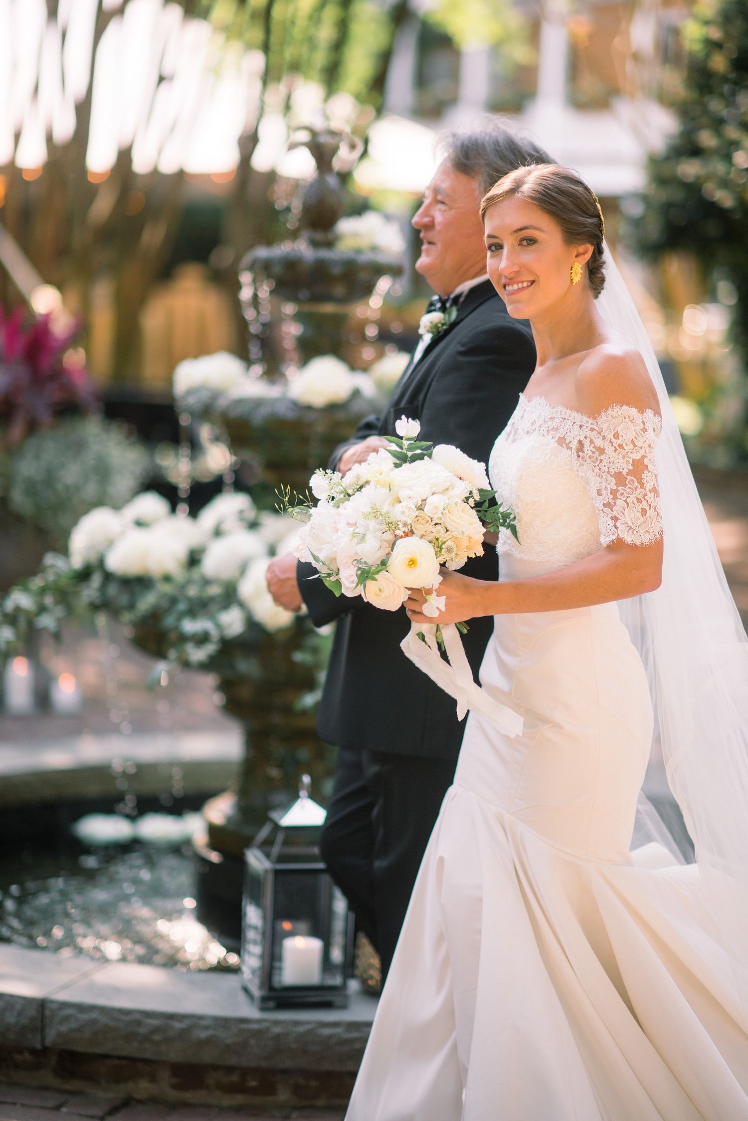 A bride in an elegant lace wedding dress holds a white floral bouquet during an outdoor ceremony.