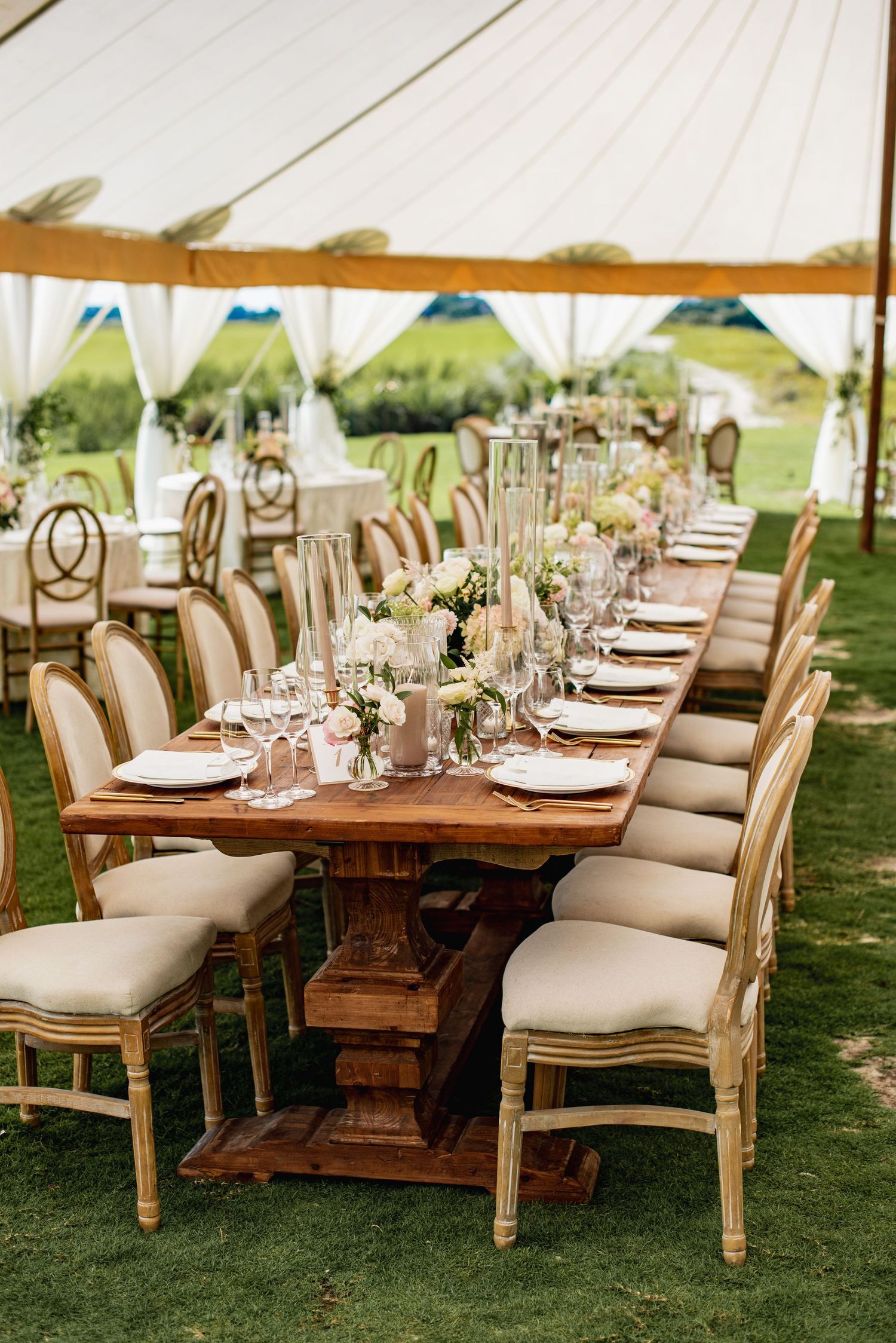 Elegant outdoor wedding reception setup with long wooden tables and upholstered chairs on a manicured lawn under white tents.