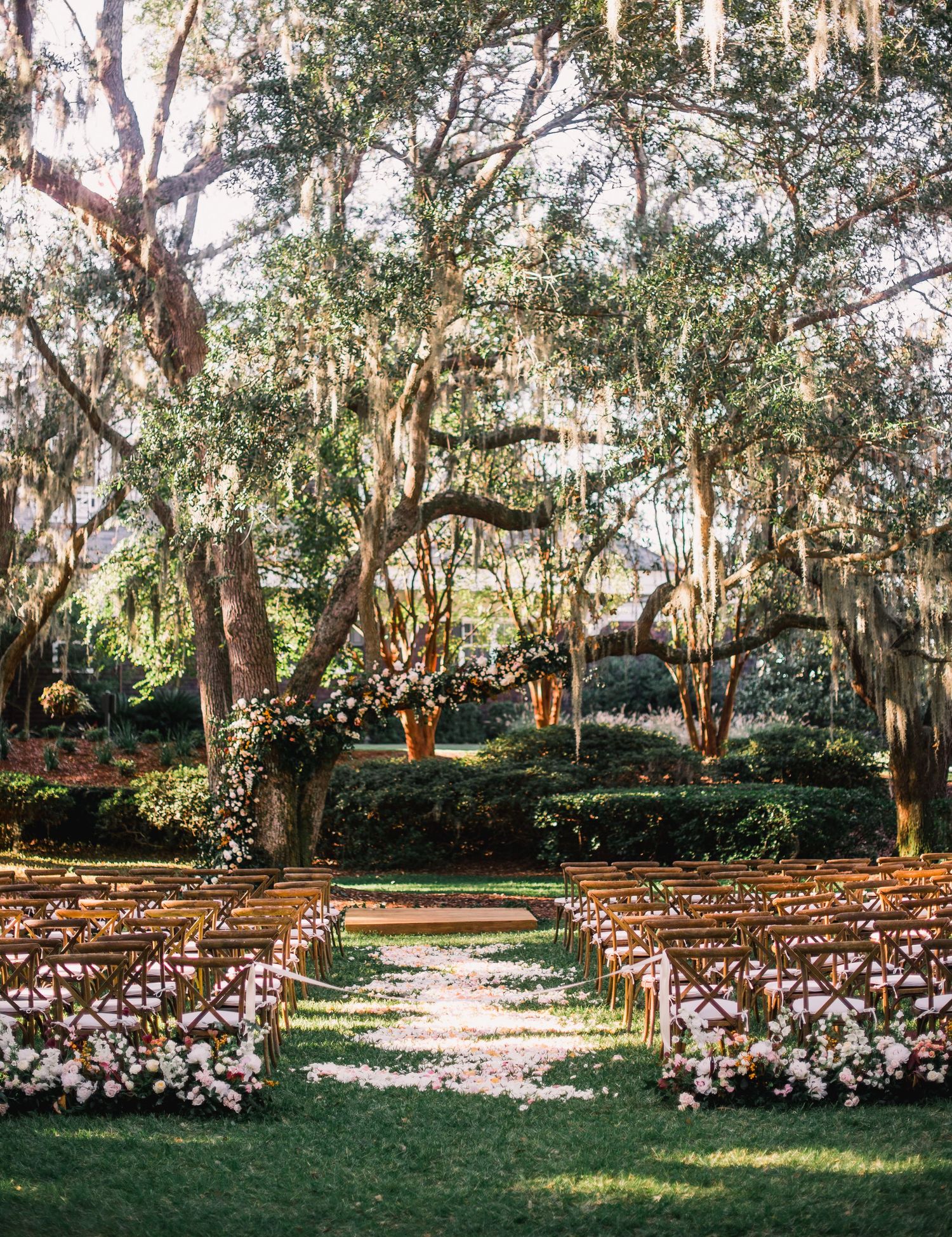 Outdoor wedding ceremony setup with rows of white chairs under Spanish moss-draped trees in a garden setting.