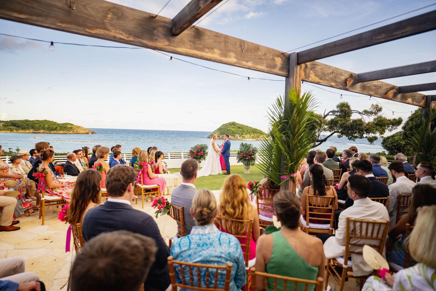 A beachfront wedding ceremony with guests seated in rows facing the ocean under a wooden pergola structure.