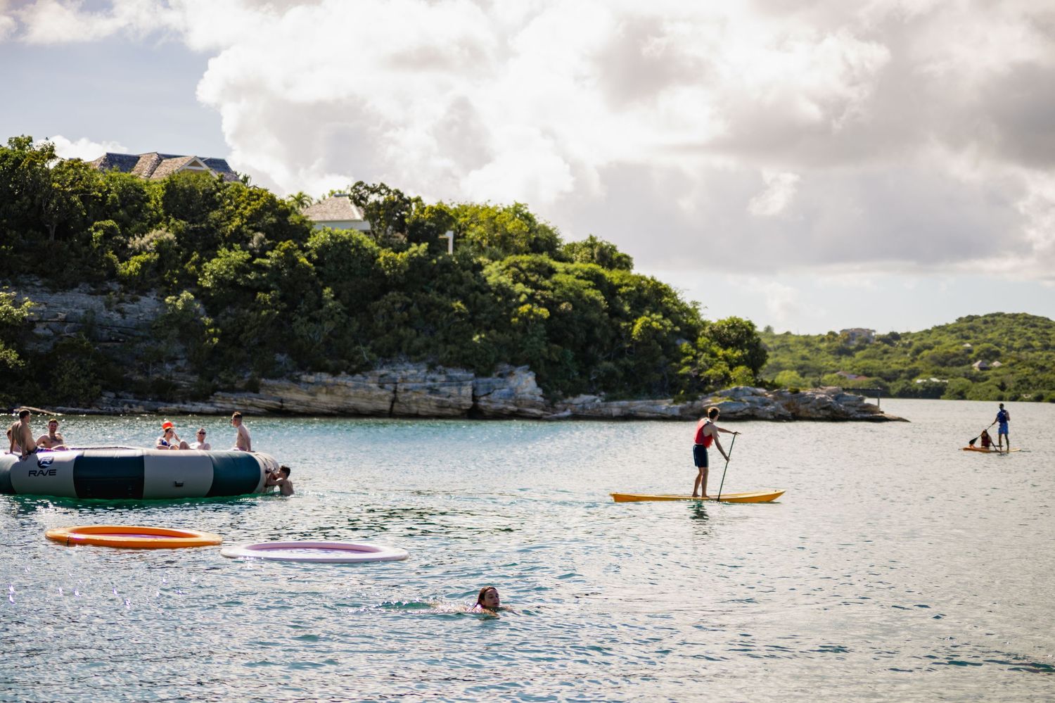 People enjoying water activities including paddleboarding and swimming at a scenic coastal cove with limestone cliffs and trees.